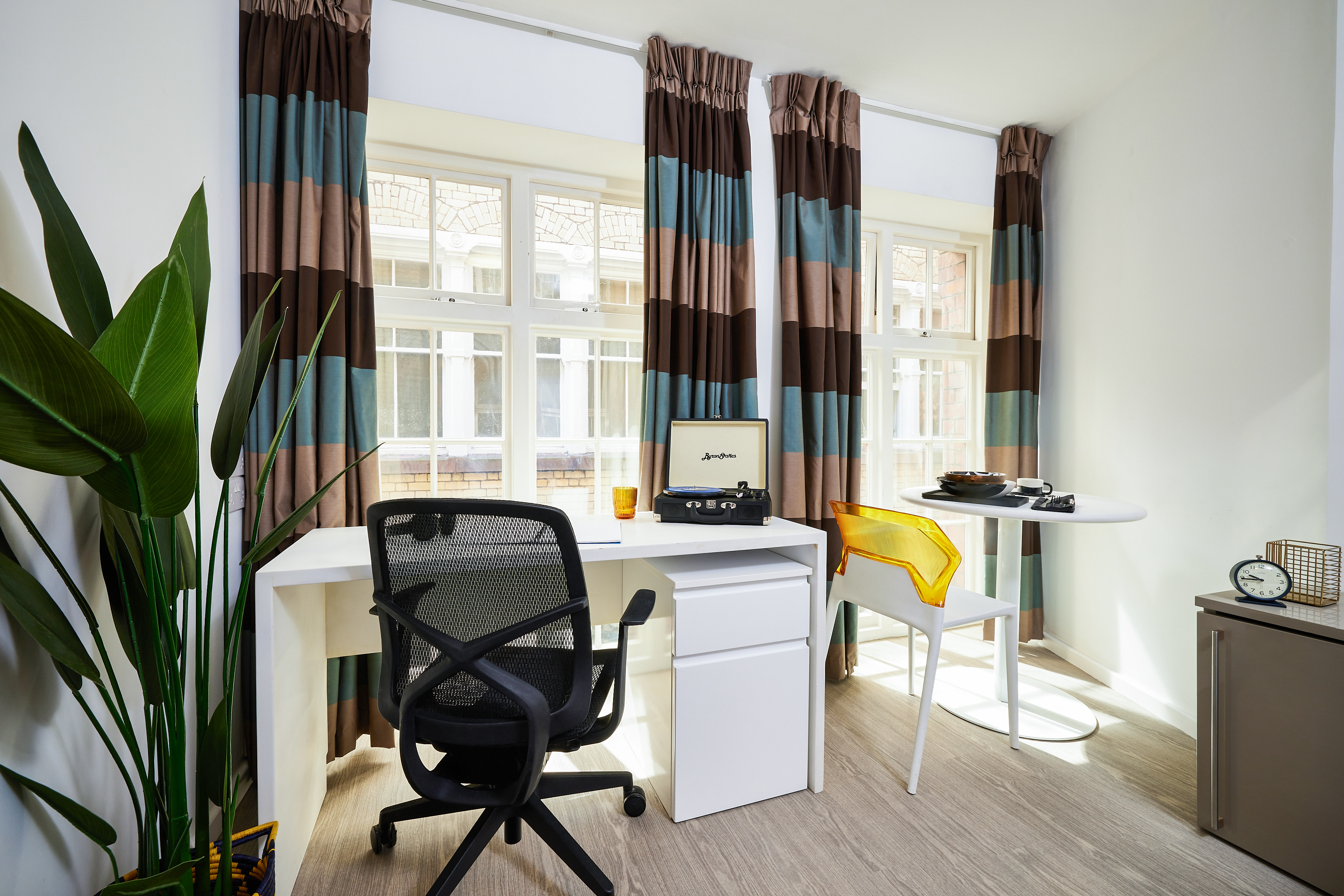 A bright workspace with a black office chair at a white desk on Crosshall Street. A record player and yellow chair are on a small round table. Large windows with striped curtains let in sunlight. A potted plant is on the left, and a clock is on a shelf.