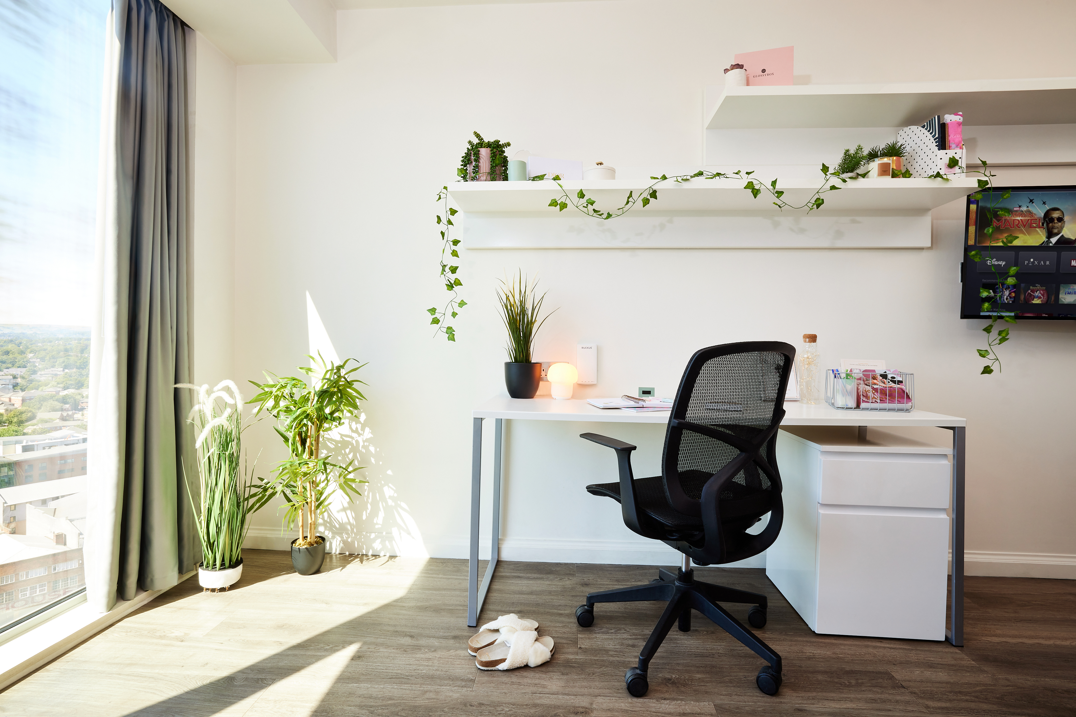 A bright, minimalist home office features a black mesh chair at a white desk topped with a lamp, potted plant, and sleek telephone. White shelves above hold greenery and decor items. A window with a city view is to the left, while plants gracefully adorn the floor.