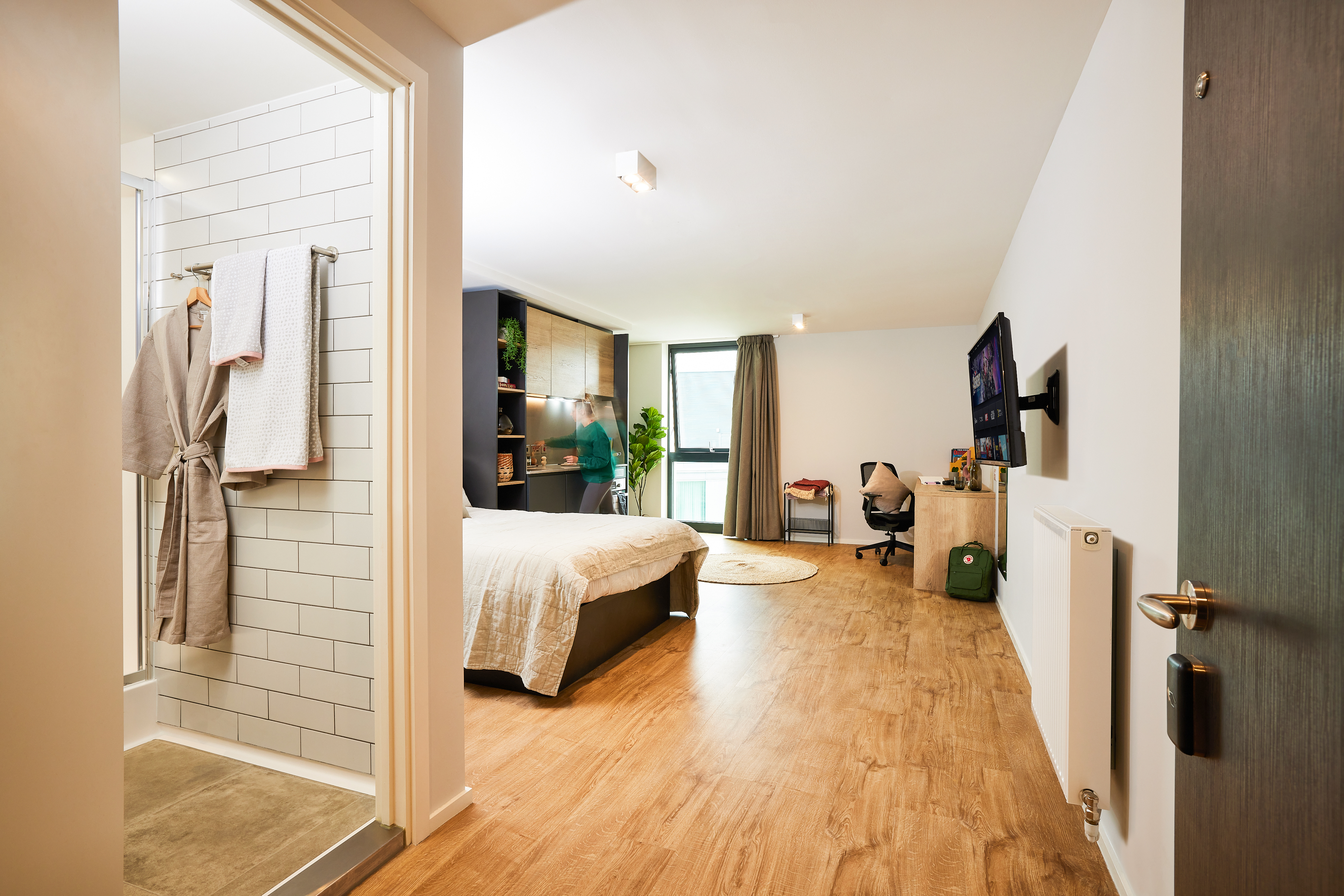 View of a modern UK bedroom with a wooden floor, featuring a neatly made bed, wall-mounted TV, desk with chair, green backpack, and window with curtains. An open bathroom is visible with a robe and towels hanging on the tiled wall.