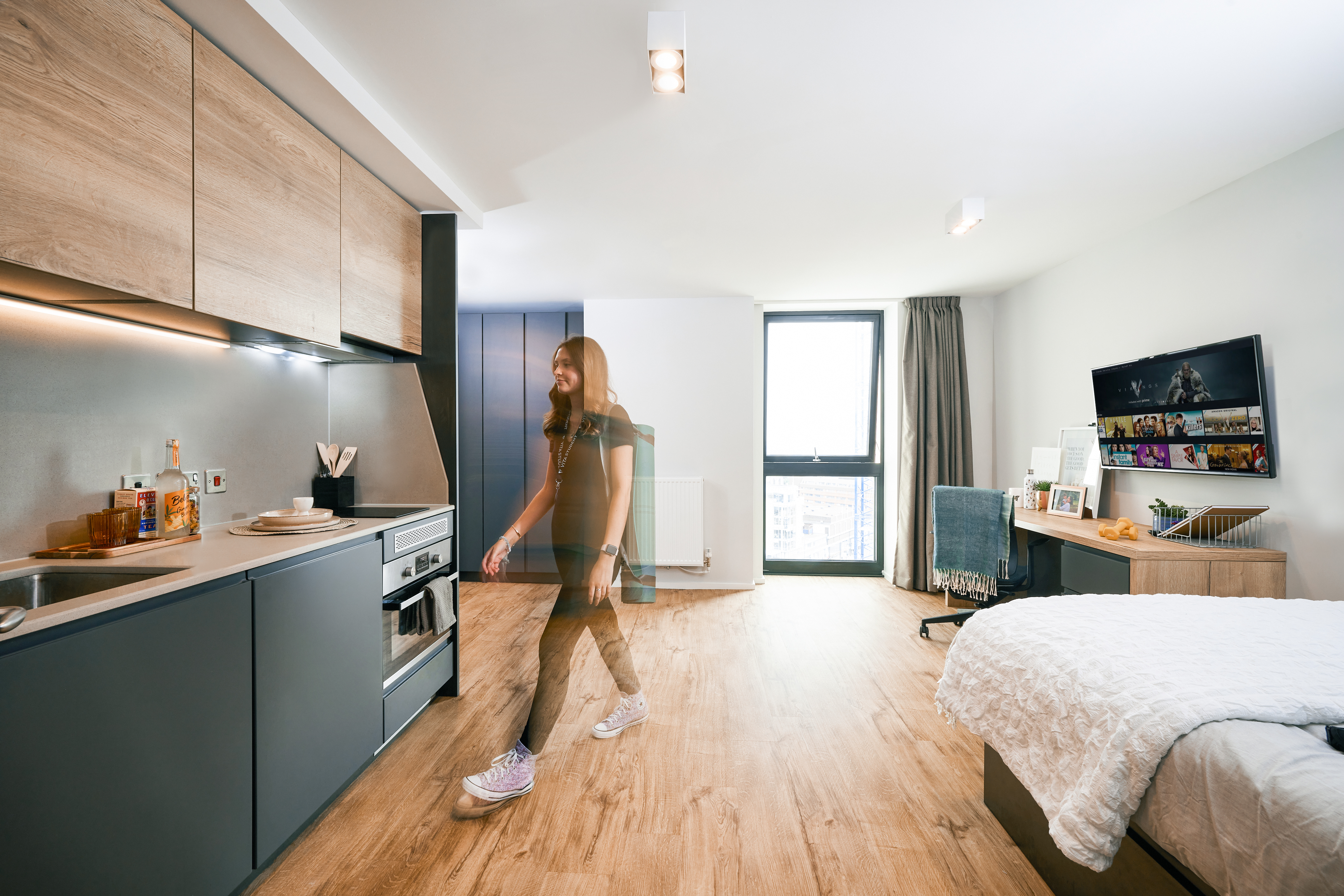 A person strolls through a modern Crescent studio apartment with a kitchen, bed, and desk area. The room features wooden flooring, a window with curtains, and a television on the wall. The kitchen boasts sleek, contemporary cabinets and appliances in true Portland style.