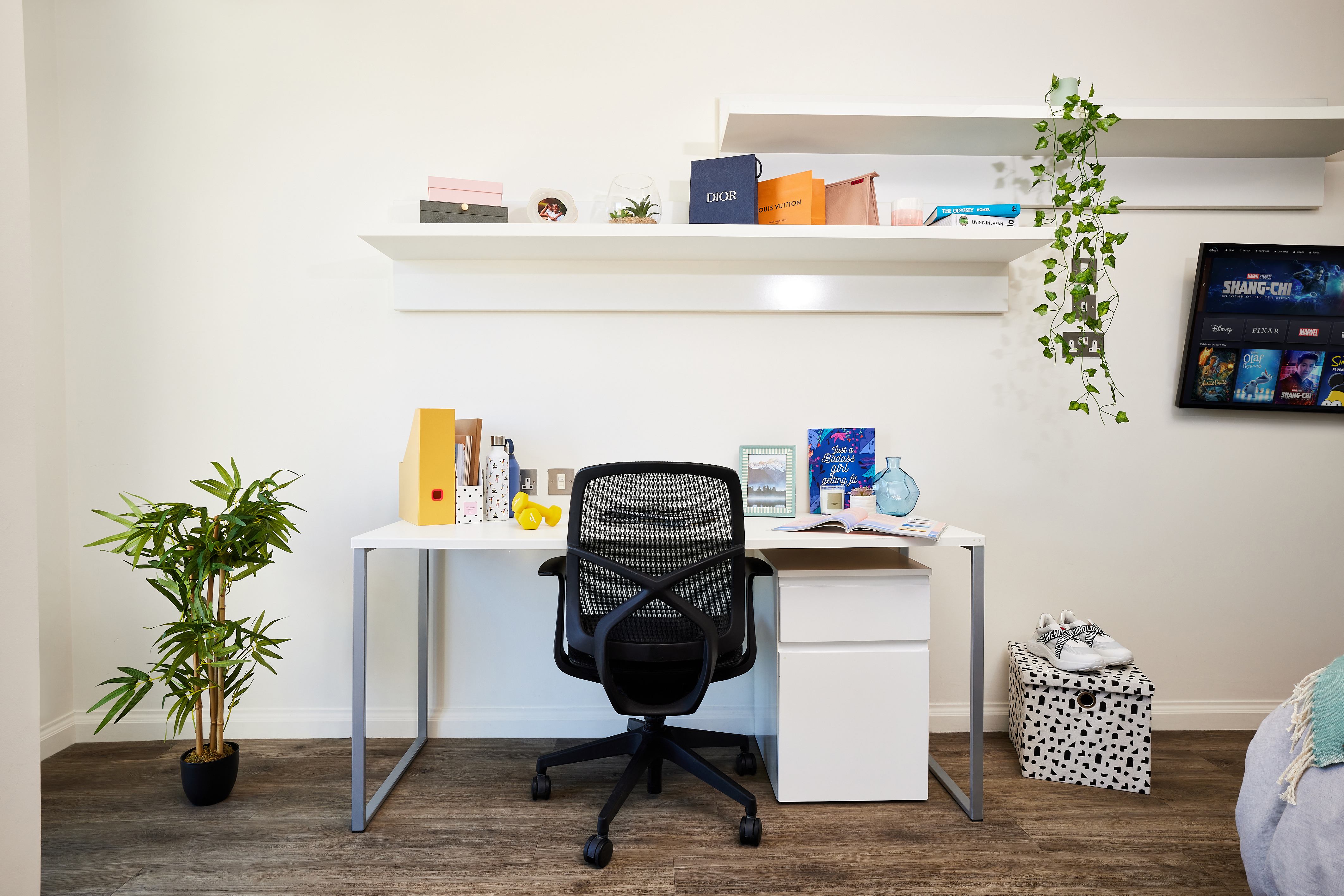 A minimalist home office features a white desk and black ergonomic chair. Shelves above hold books and plants, while a small indoor plant sits beside the desk. A tablet displaying movie posters is mounted on the wall, reminiscent of a classic Telephone House aesthetic. A storage box rests on the floor.