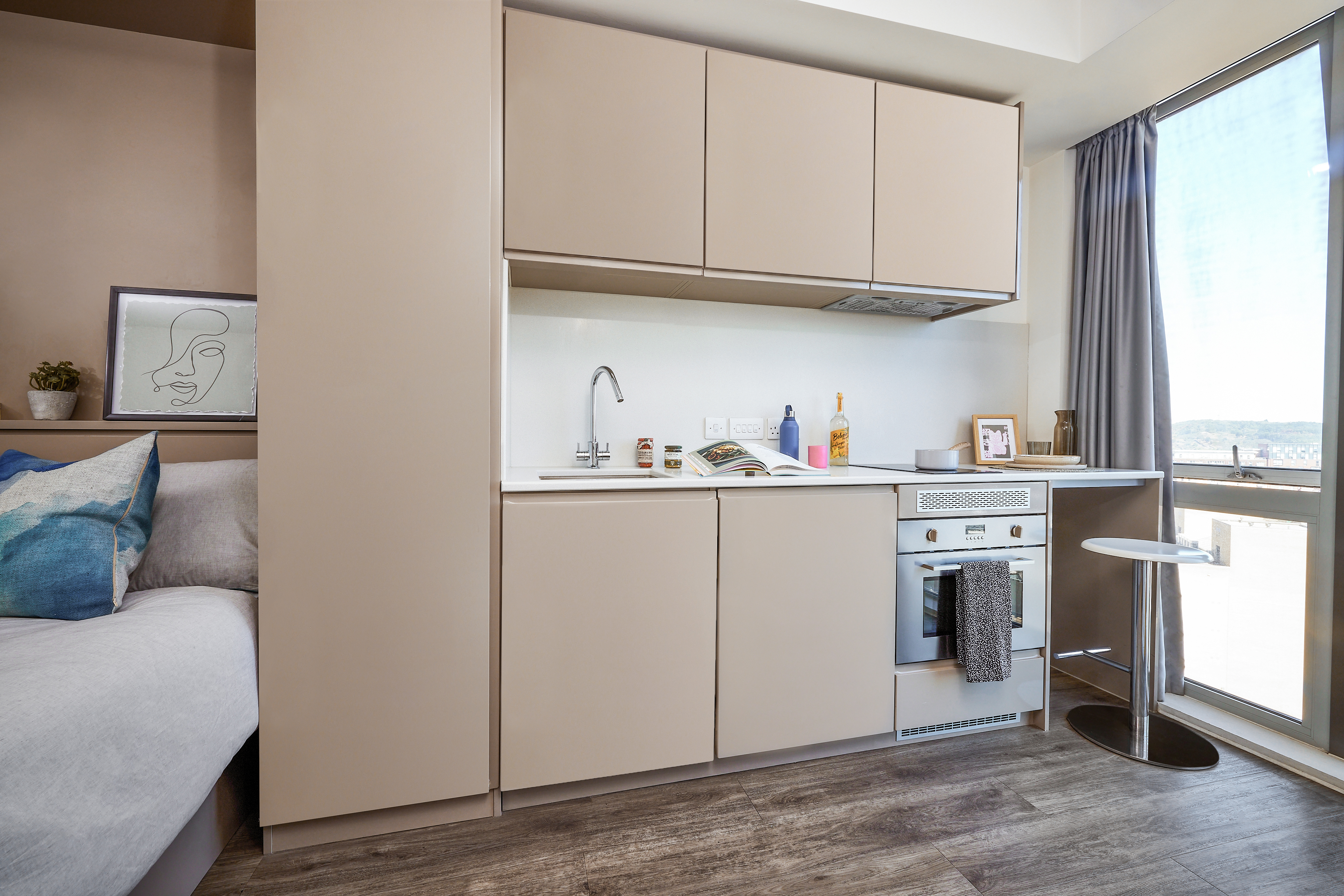 In a cozy studio apartment, the small modern kitchen features beige cabinets and a sleek countertop with a sink, stove, and oven. By the large window with a sheer curtain sits a round table with a stool. Nearby, minimalistic artwork accompanies vintage house-themed decor pieces.