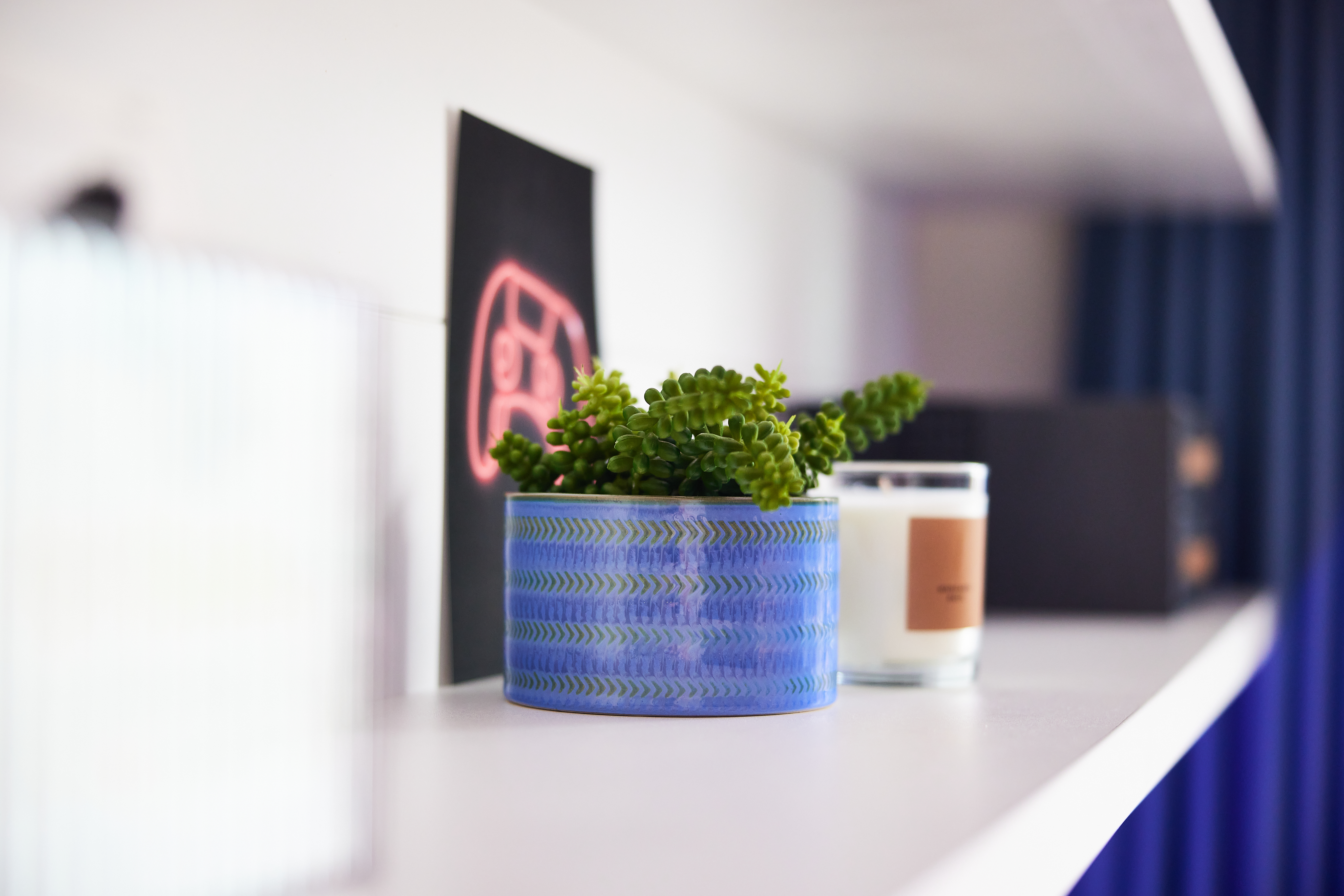 A close-up of a shelf at Portland House showcases a blue textured pot with a succulent plant, alongside a small candle adorned with a brown label. In the background, blurred yet distinct, lies black artwork featuring a pink design.