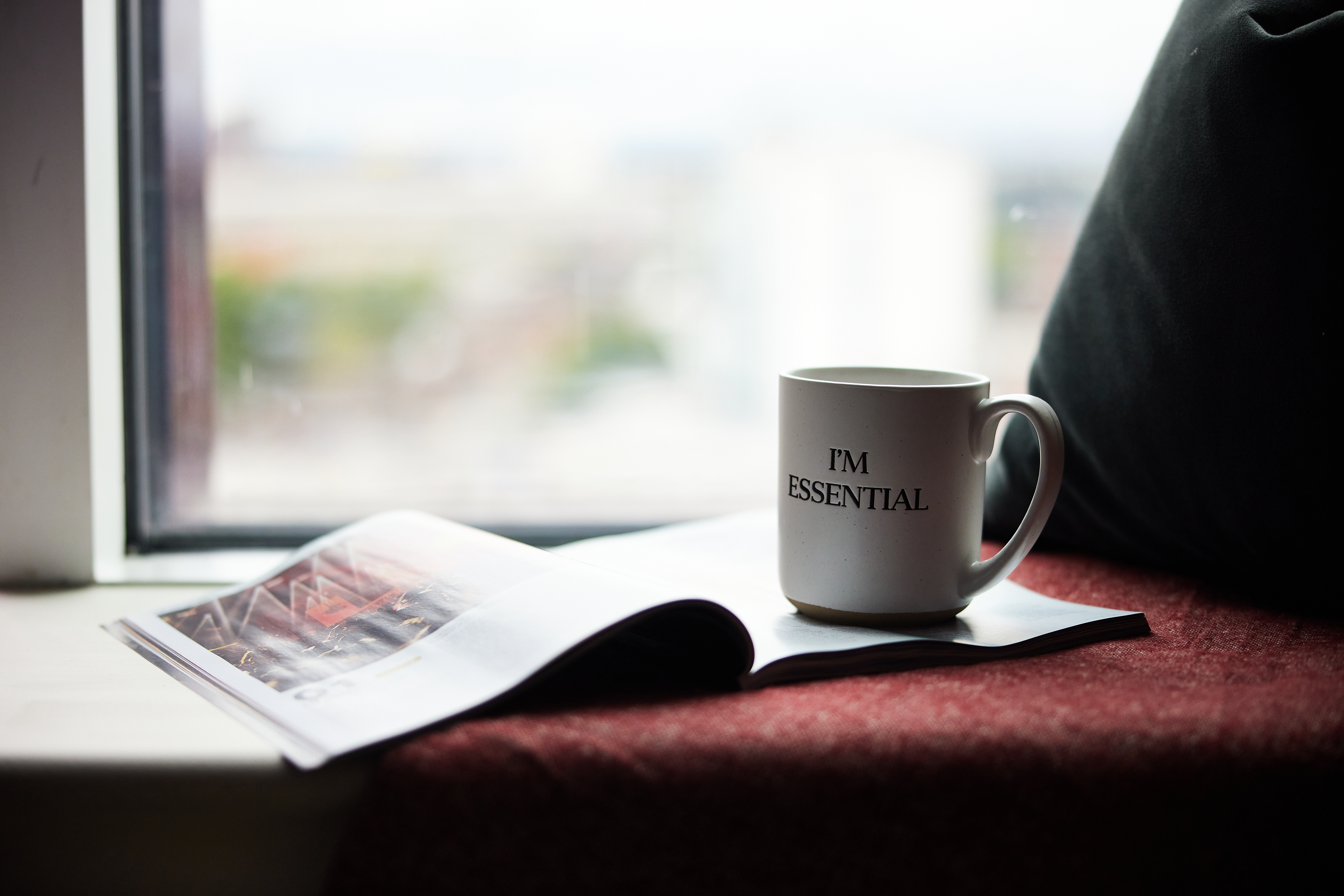 A white mug with "I'M ESSENTIAL" text sits on an open magazine, nestled on a red cushioned windowsill of a cozy Portland house. Natural light streams in through the window, illuminating the scene. A dark pillow is placed beside the magazine.