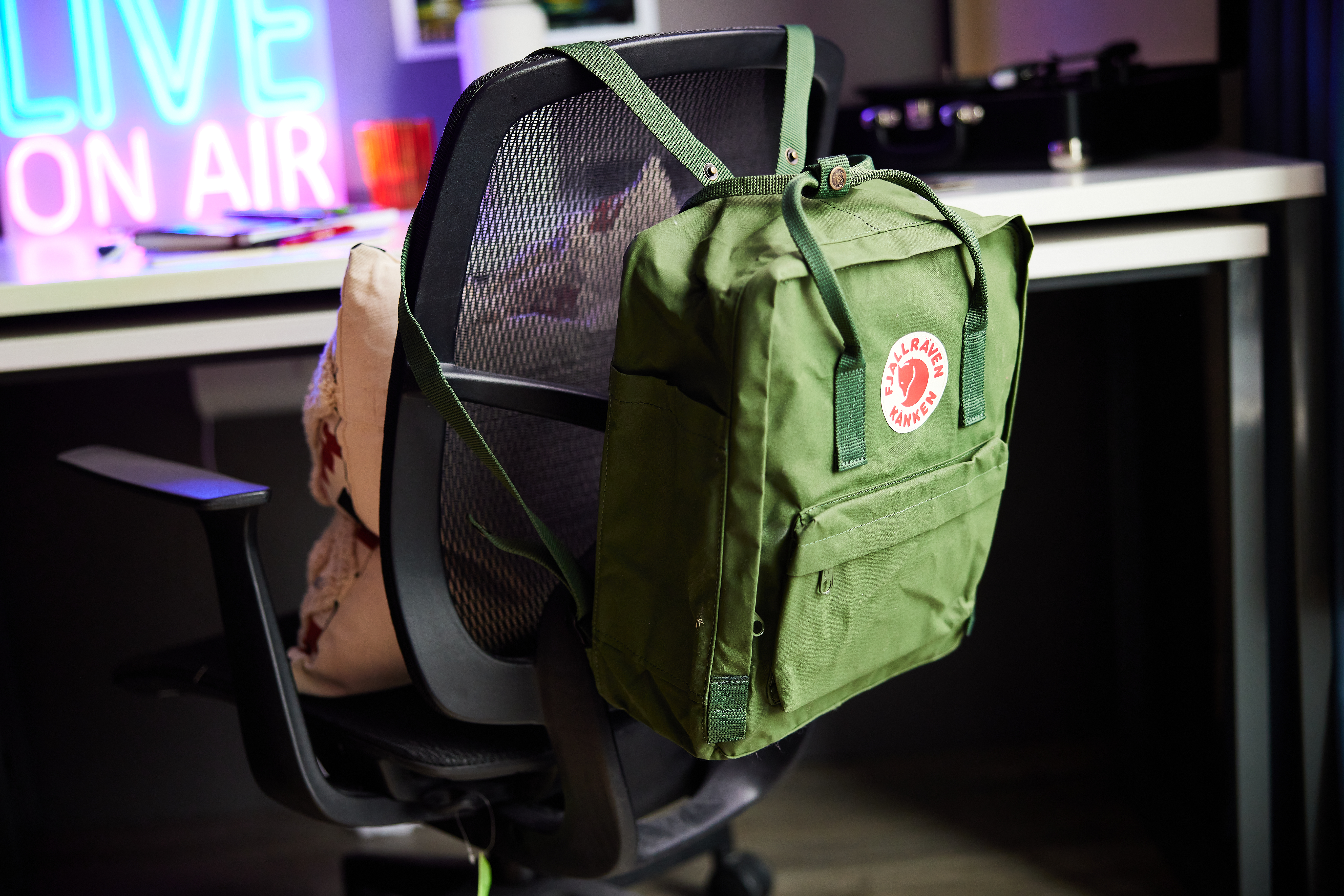 A green backpack with a circular patch hangs on a black office chair at Portland House. Behind the chair, a desk features illuminated signage reading "LIVE ON AIR" in blue and pink neon lights. A pillow is also placed on the chair.
