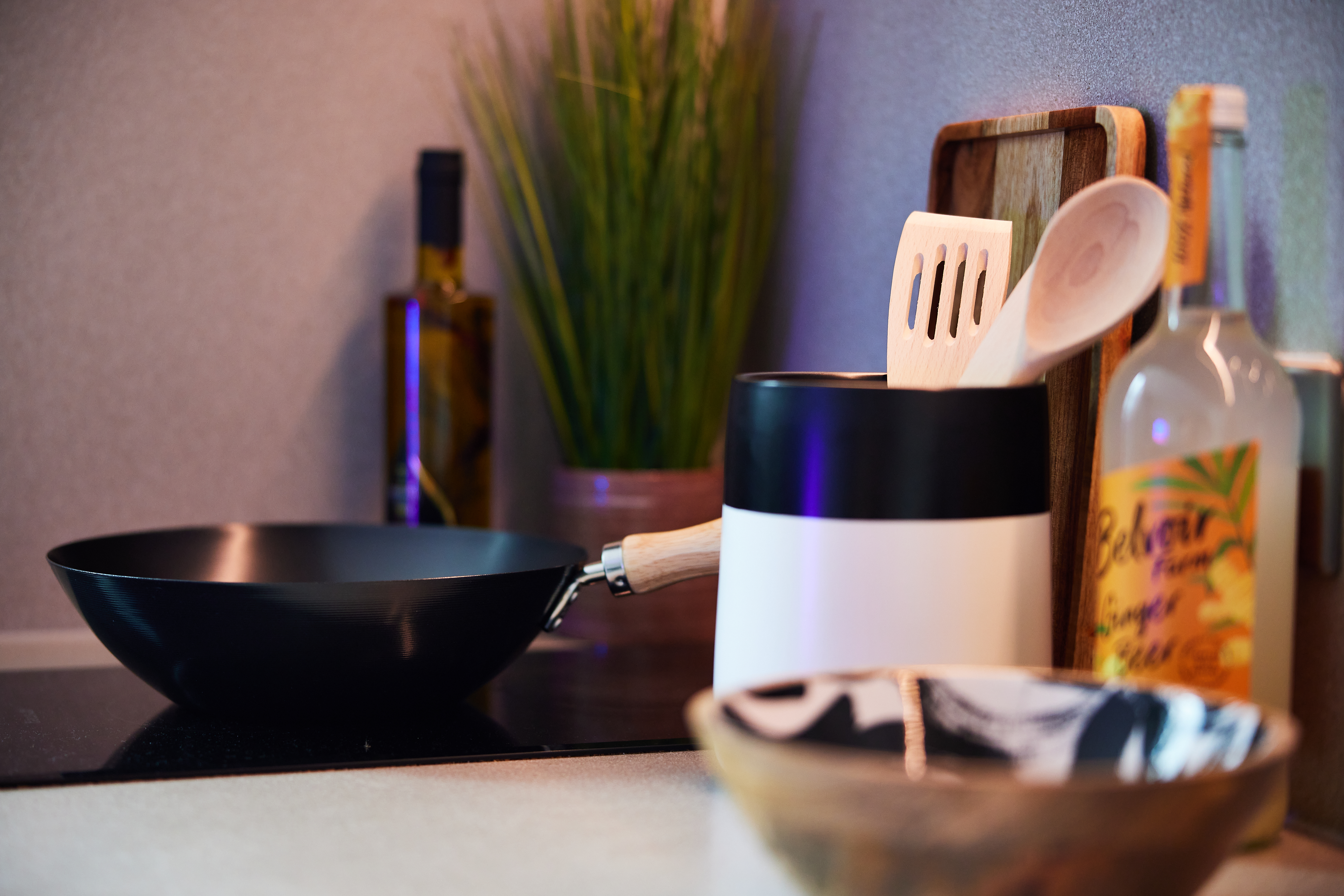 In a cozy Portland House kitchen scene, a black frying pan sits on the stove beside a container brimming with utensils. A wooden cutting board rests nearby, accompanied by a potted plant and bottles of oil and vinegar. In the foreground, a decorative bowl adds charm to the setting.