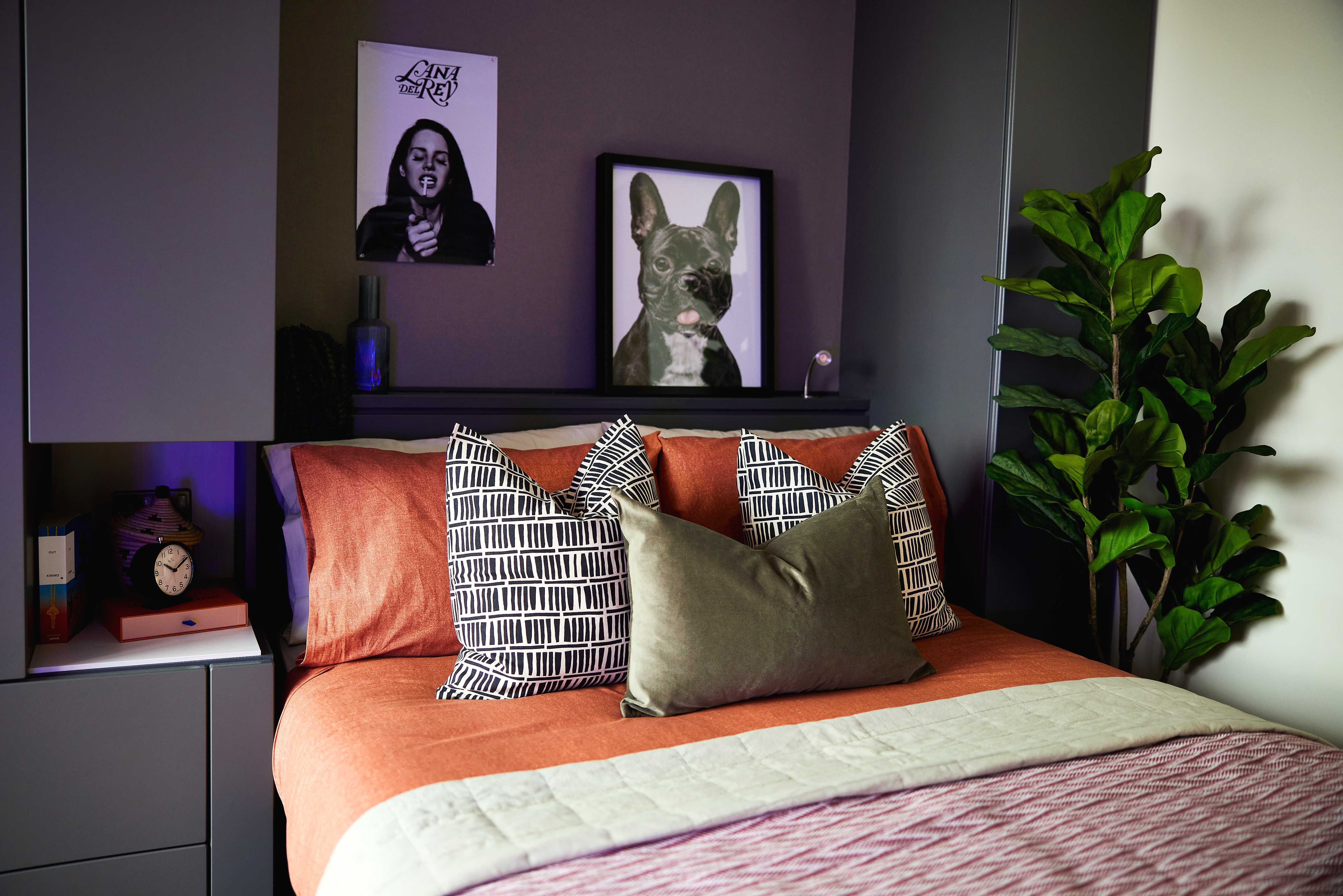 A cozy bedroom with a neatly made bed featuring rust and green pillows. Two square-framed art pieces hang above: a black and white photo of a woman and a painting of a French bulldog. A potted plant stands in perfect circle symmetry to the right of the bed.