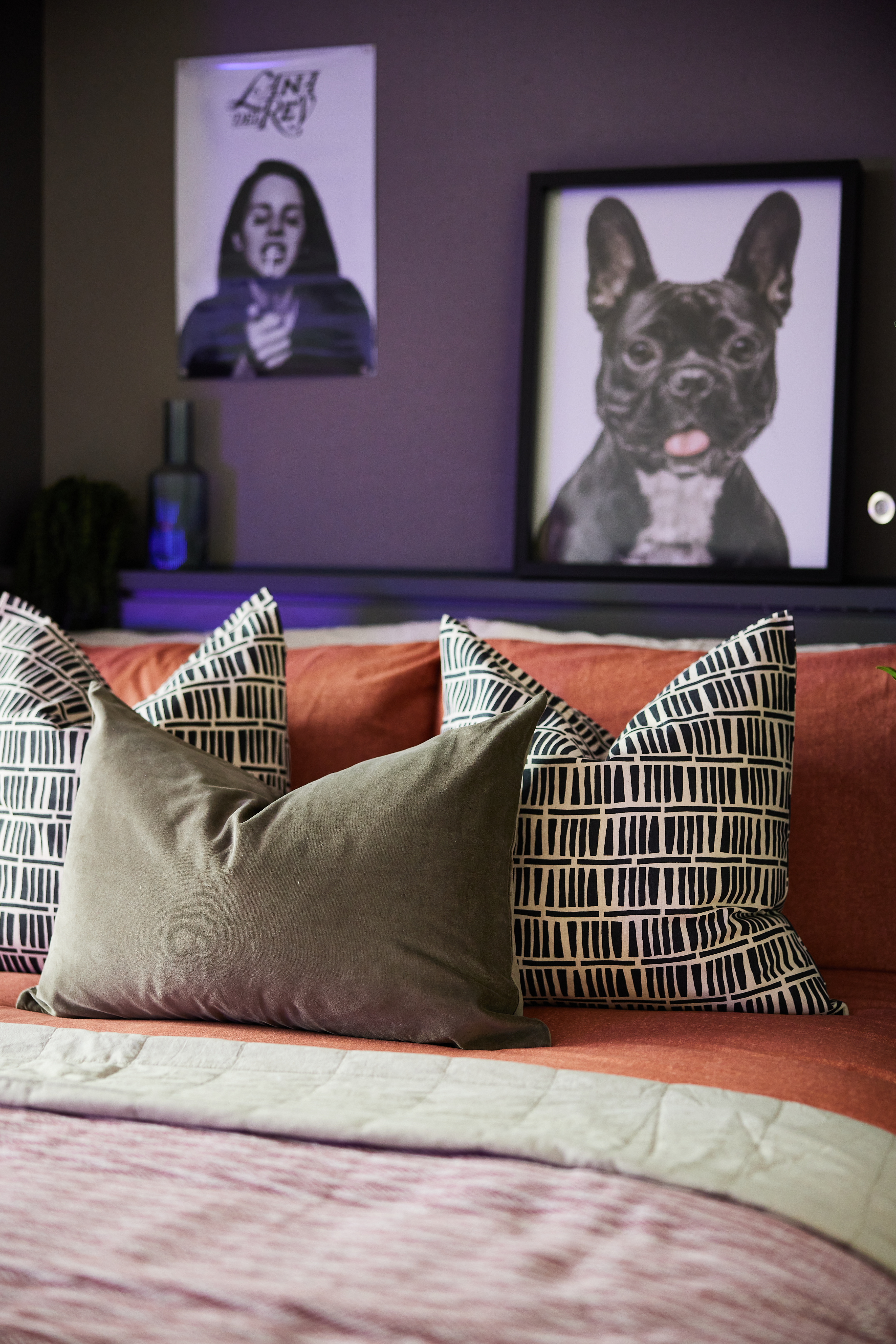 A cozy bedroom in the Portland House features a bed with patterned pillows and a solid green cushion. Above the bed, two framed pictures hang on the wall: one of a smiling French Bulldog and another with an image of a musician.