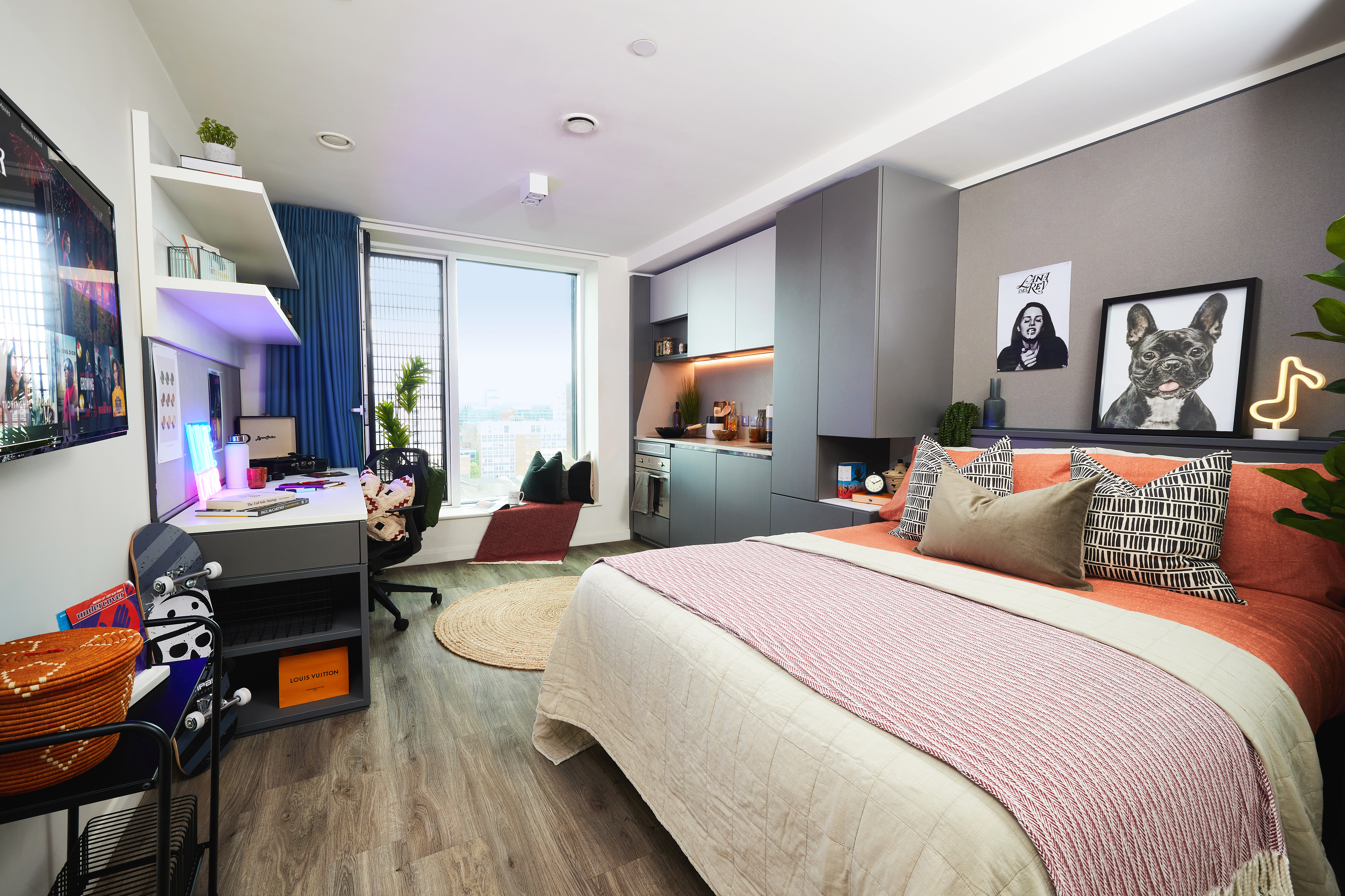 A modern, cozy bedroom features a double bed adorned with pillows and a red throw. By the window with its city view sits a desk, complete with a computer and chair. Shelves hold plants and decor in square frames, while circular art prints of a cat adorn the walls. A small kitchen area is visible.
