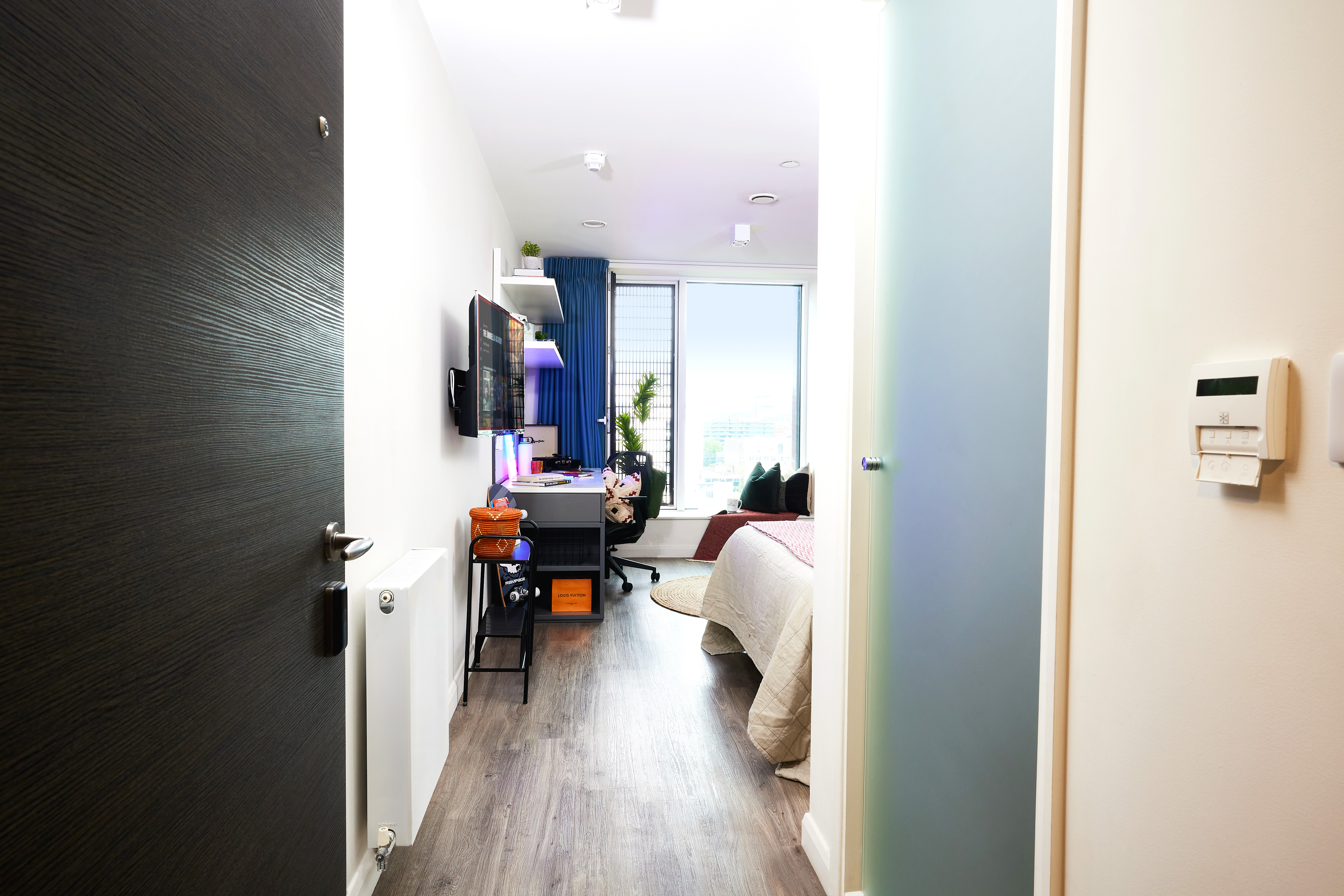 View into a bright, modern bedroom. A bed is on the right, with a desk and computer setup near a window at the back. A circular rug adds charm to the dark wood-paneled floor. A chair is near the desk, and various decorations are around the room.