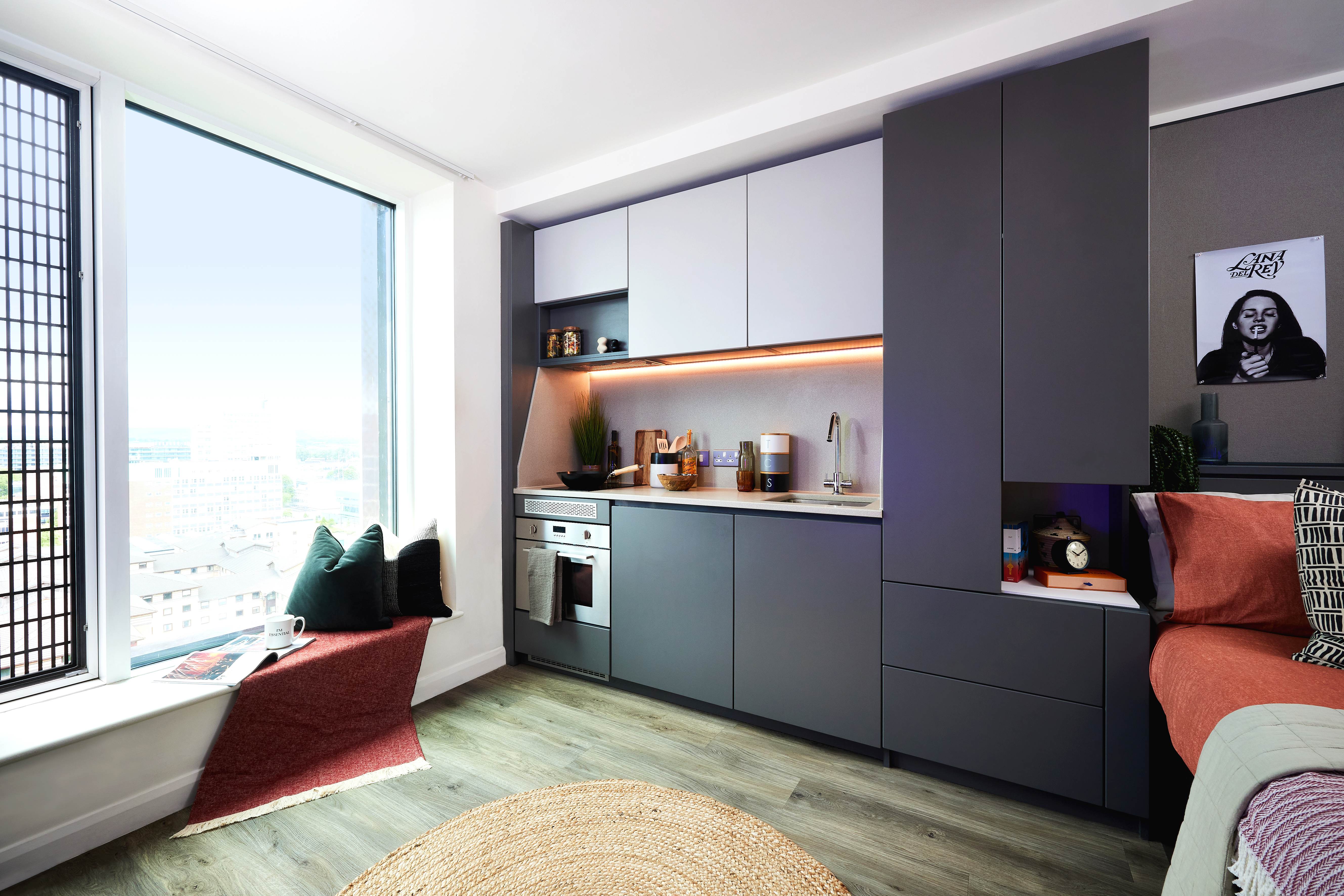Modern studio kitchen interior with sleek gray cabinets and under-cabinet lighting. A built-in oven and sink are visible. A large window with a cozy nook and cushions offers city views. Circular rug on the wood floor echoes the clean lines of the square pillows on the bed to the right.