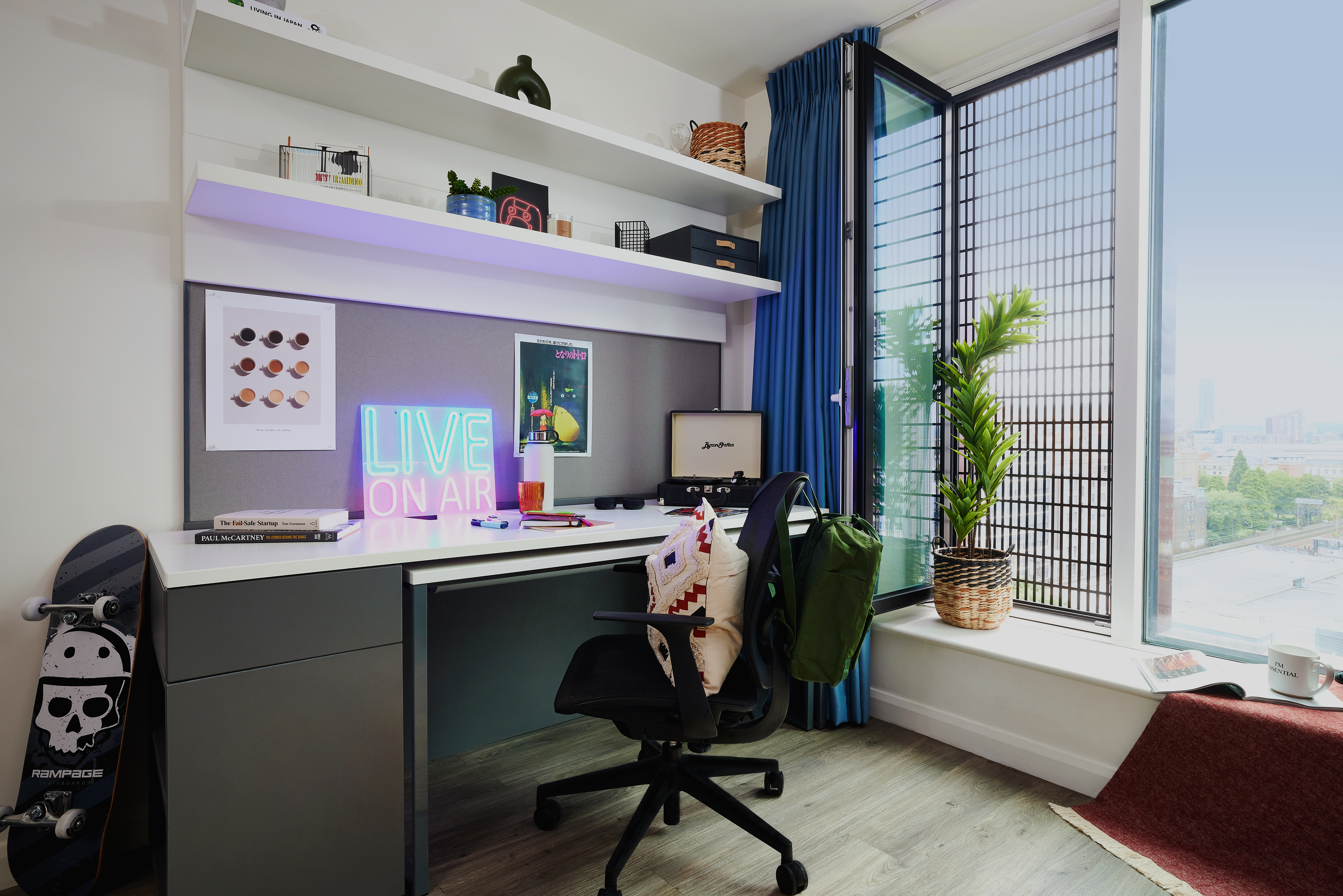 Modern workspace with a square desk, computer, and chair. Decor includes a skateboard, circle-patterned poster, and "LIVE ON AIR" neon sign. Large window with city view, small indoor plant, and shelves with books. Blue curtains partially drawn over light wood floor.