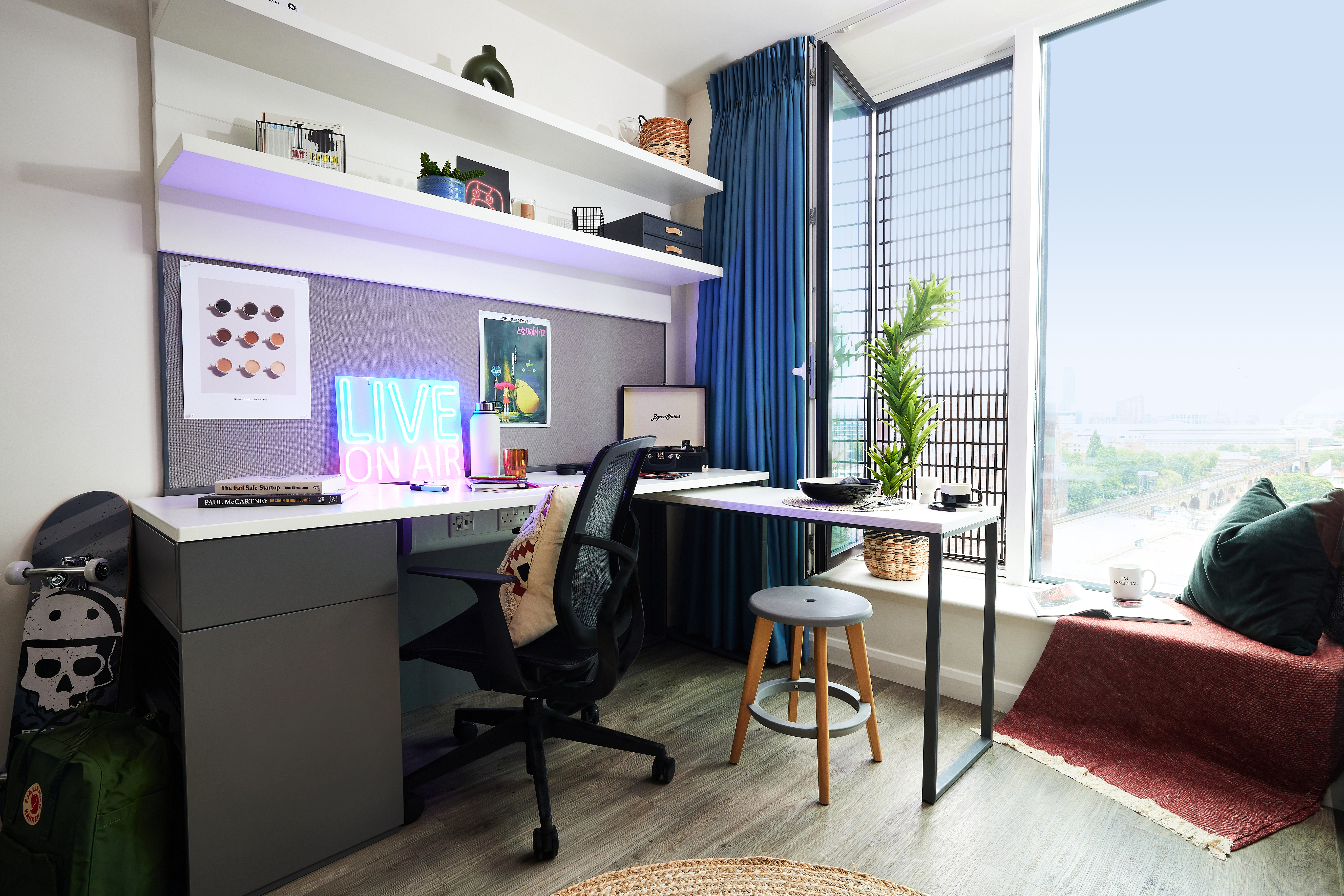 A modern home office features a sleek desk, chair, and laptop. A "LIVE ON AIR" neon sign hangs on the wall next to a circular clock. A skateboard and backpack rest near a square cabinet. Blue curtains frame the large window with city views, while a plant and red throw blanket add warmth.