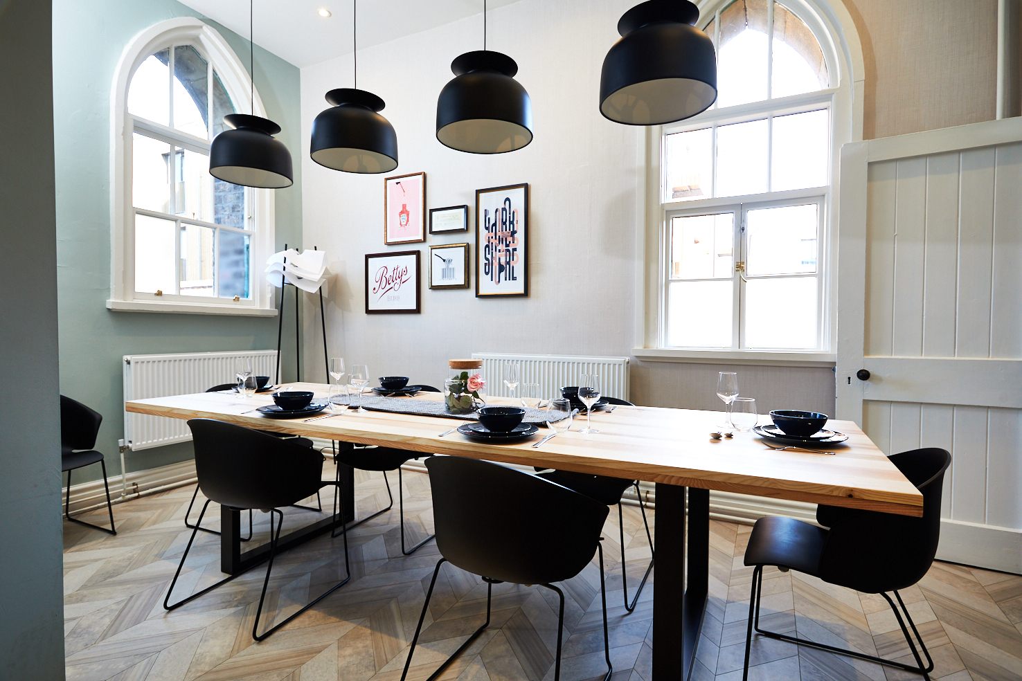 A modern dining room with a long wooden table, six black chairs, and black pendant lights. The walls have large arched windows and framed artwork. The floor features a herringbone pattern. A white floor lamp stands in the corner.