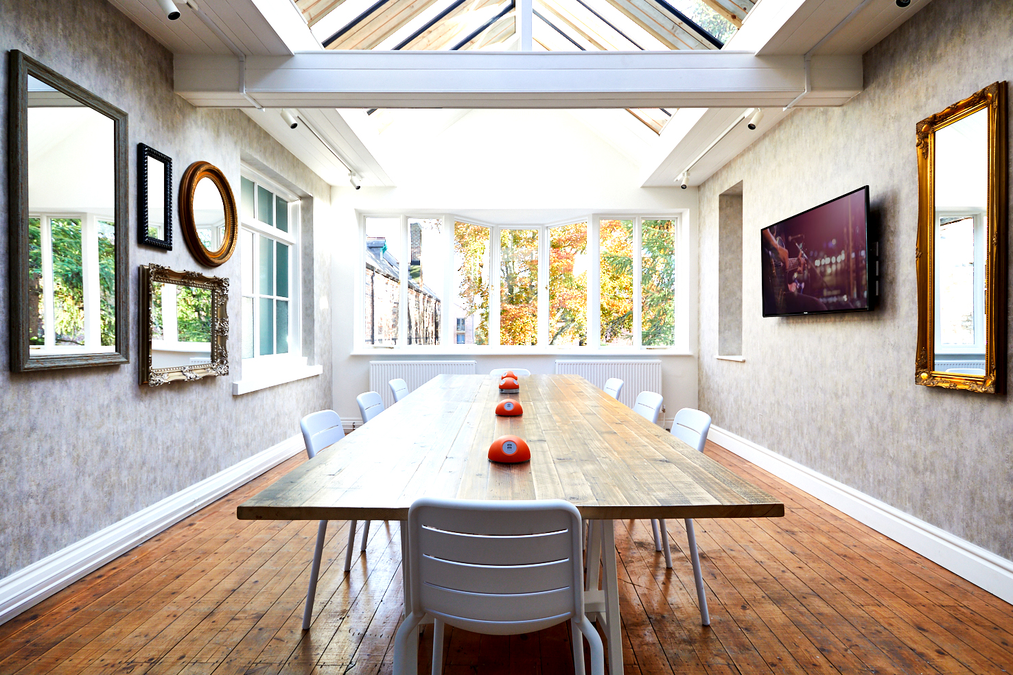 Set on Lawrence Street, this spacious, well-lit conference room features a long wooden table paired with white chairs. The walls are adorned with assorted mirrors and a TV, while skylights and large windows flood the space with light. Orange bowl centerpieces add a touch of warmth along the table.