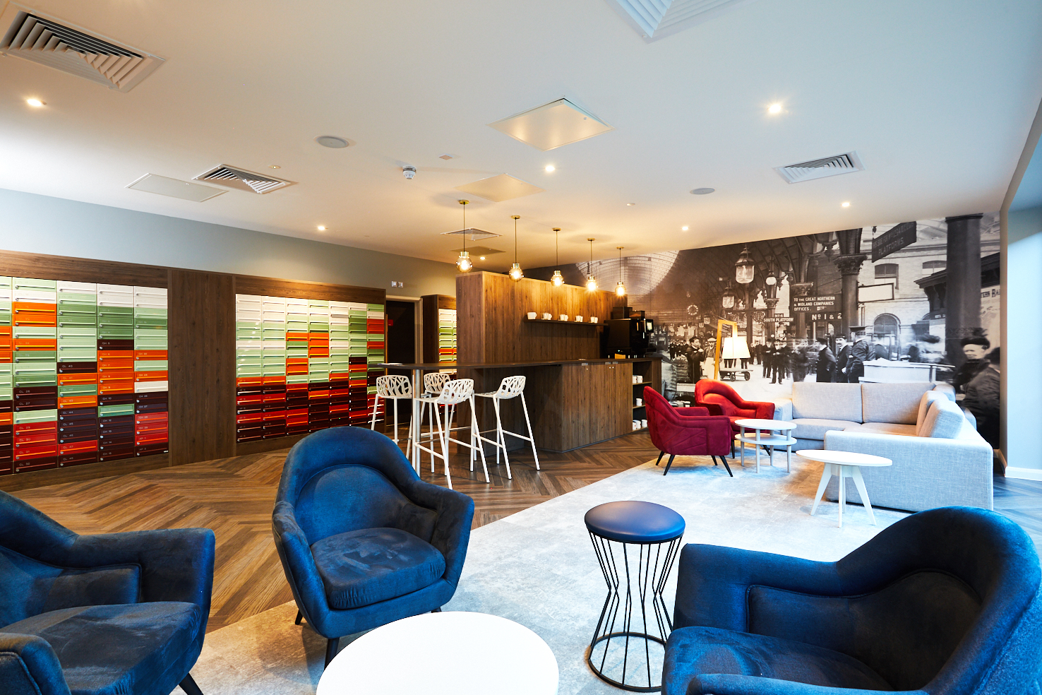 Modern lounge area with colorful lockers on one wall and a large vintage photo mural on the other. Features include cozy blue and red chairs, tall bar stools by a wooden counter, and a sofa set on a light rug. Soft lighting enhances the warm atmosphere.