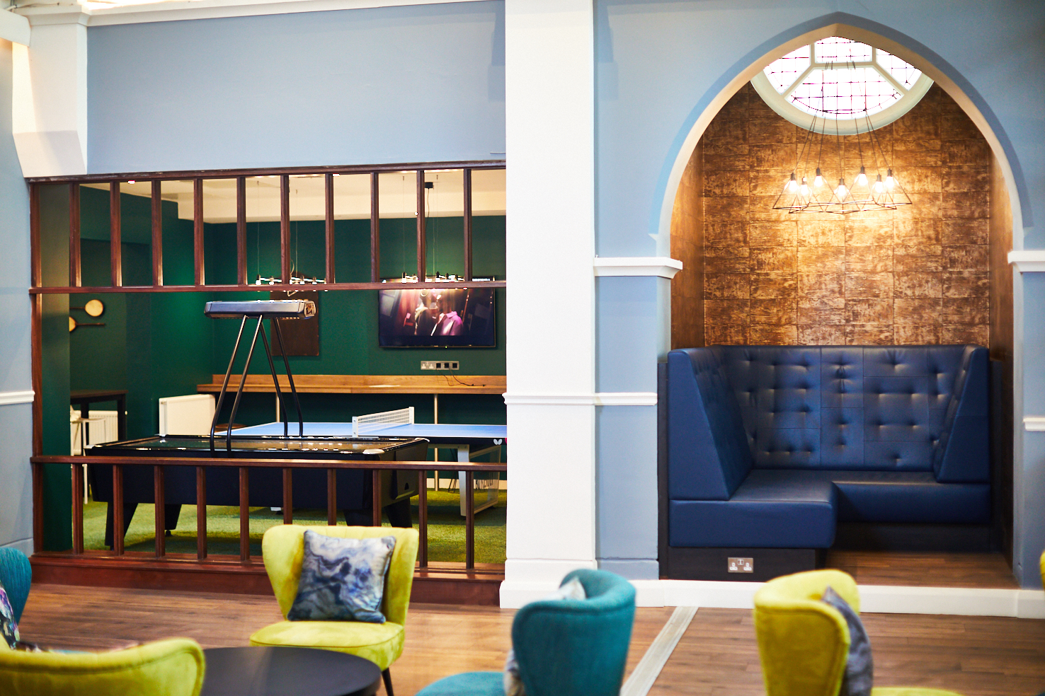A stylish lounge on Lawrence Street features a pool table under a light fixture, a wall-mounted TV, and a cozy, arched alcove with blue cushioned seating. Colorful chairs and a round table sit in the foreground against the vibrant blue and green wall backdrop.