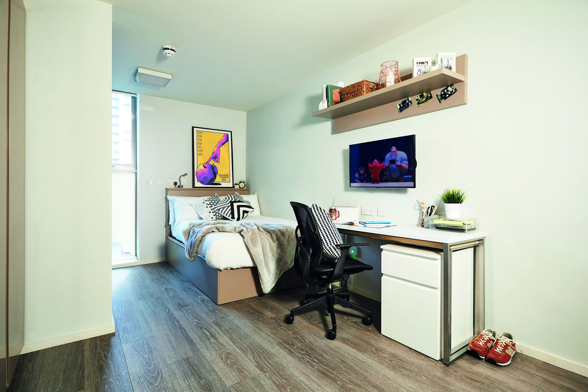 A tidy bedroom with a bed on the left and a desk set up like an office building workspace on the right. A TV is mounted on the wall, with a shelf above holding books and decorations. Light floods in through a large window at Telephone House, where sneakers rest near the desk.