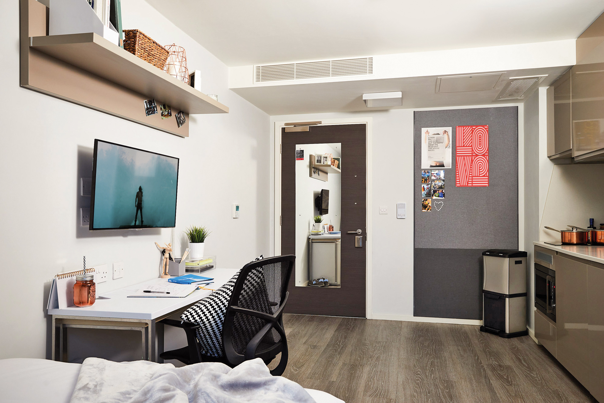 A modern dorm room in Telephone House features a desk and chair facing a wall-mounted TV. The desk holds books, a lamp, and stationery. Shelves and a bulletin board with photos and a poster adorn the walls. A small fridge sits in the corner near the door, illuminated by soft lighting.