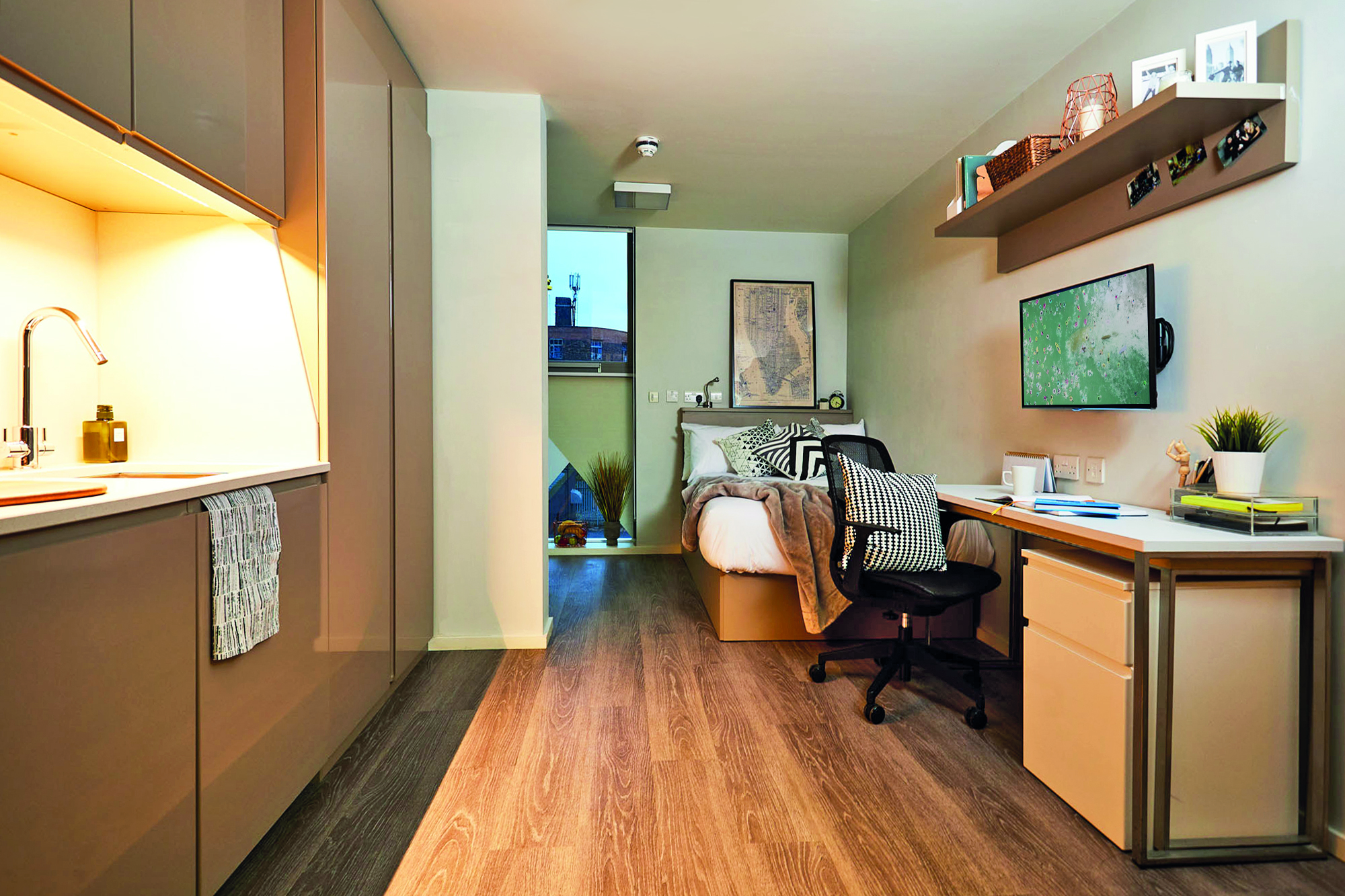 Modern studio apartment with a bed, desk, and chair. A small kitchenette is on the left, and a wall-mounted TV is across from the bed. Shelves hold books and decor. A window at the end provides natural light. Neutral tones dominate this cozy house retreat, complete with efficient design.