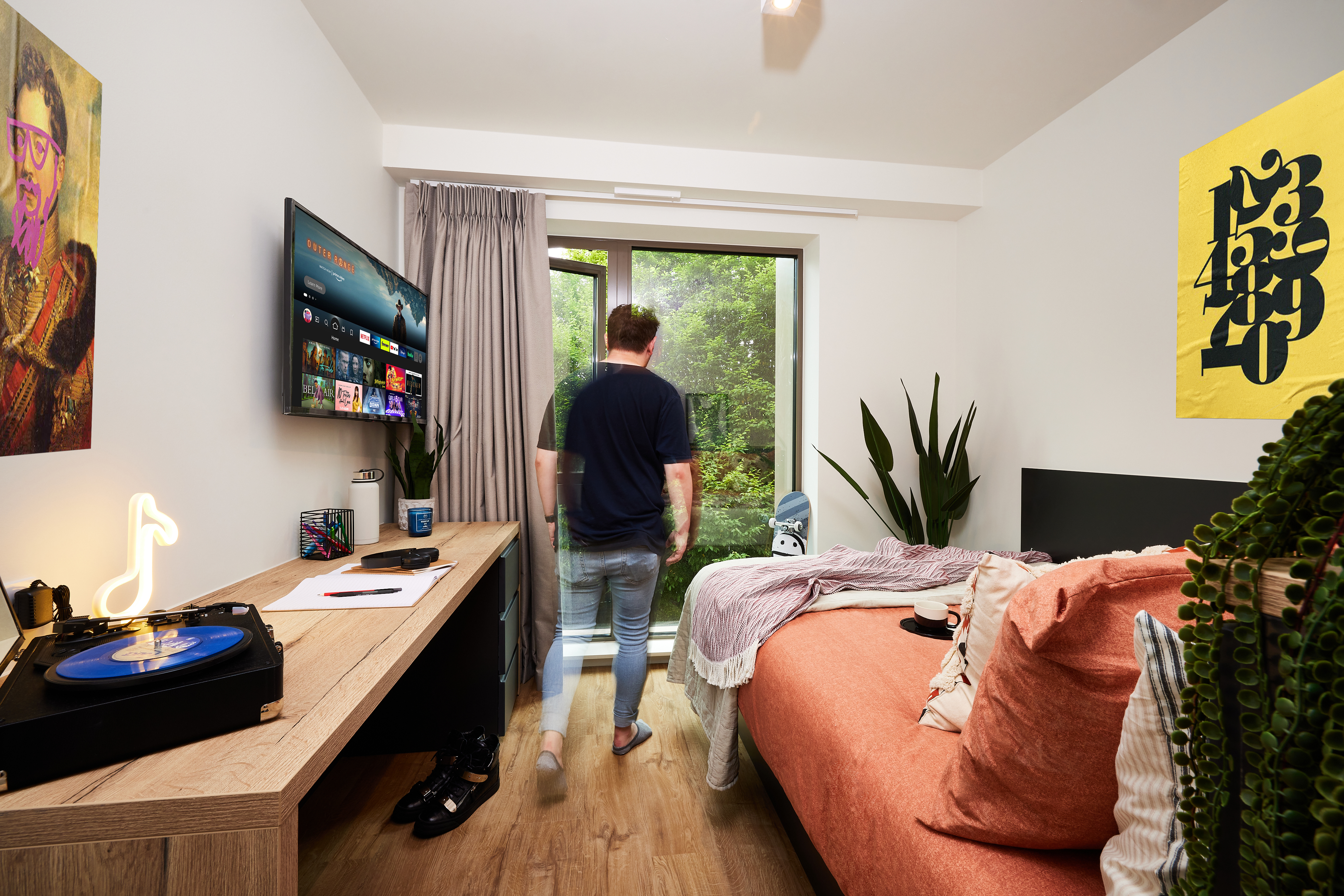 A person stands in a small, modern bedroom in Cannon Park, with wood flooring, looking out the glass door at greenery. The room has a bed, desk with a neon light and TV, turntable, abstract art, and several plants.