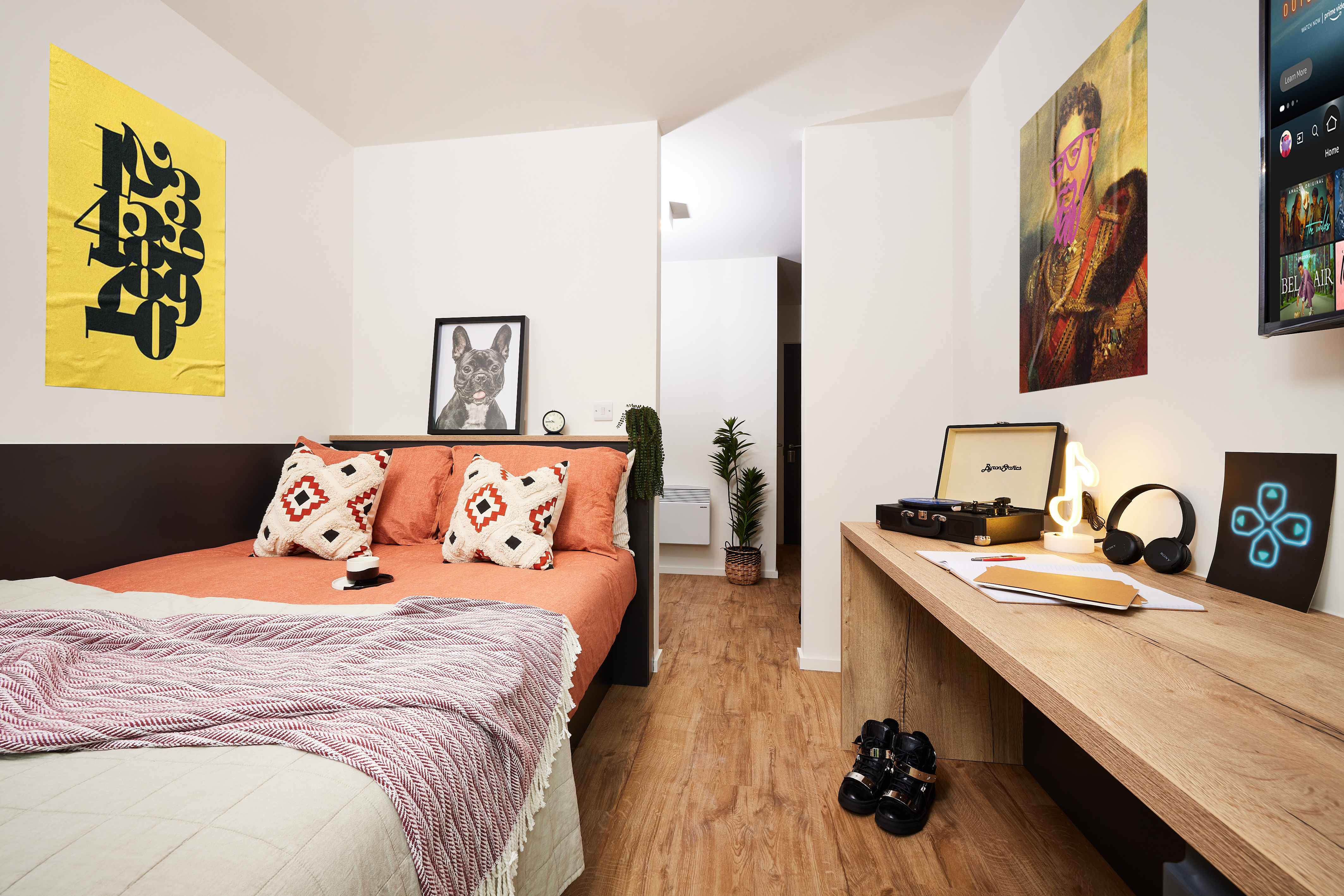 A cozy bedroom in Warwick features a bed with orange and beige linens. A record player and headphones sit on a wooden desk. The walls display colorful artwork, while a TV and potted plant add charm. The wooden floor leads to a hallway that opens towards the scenic views of Cannon Park.