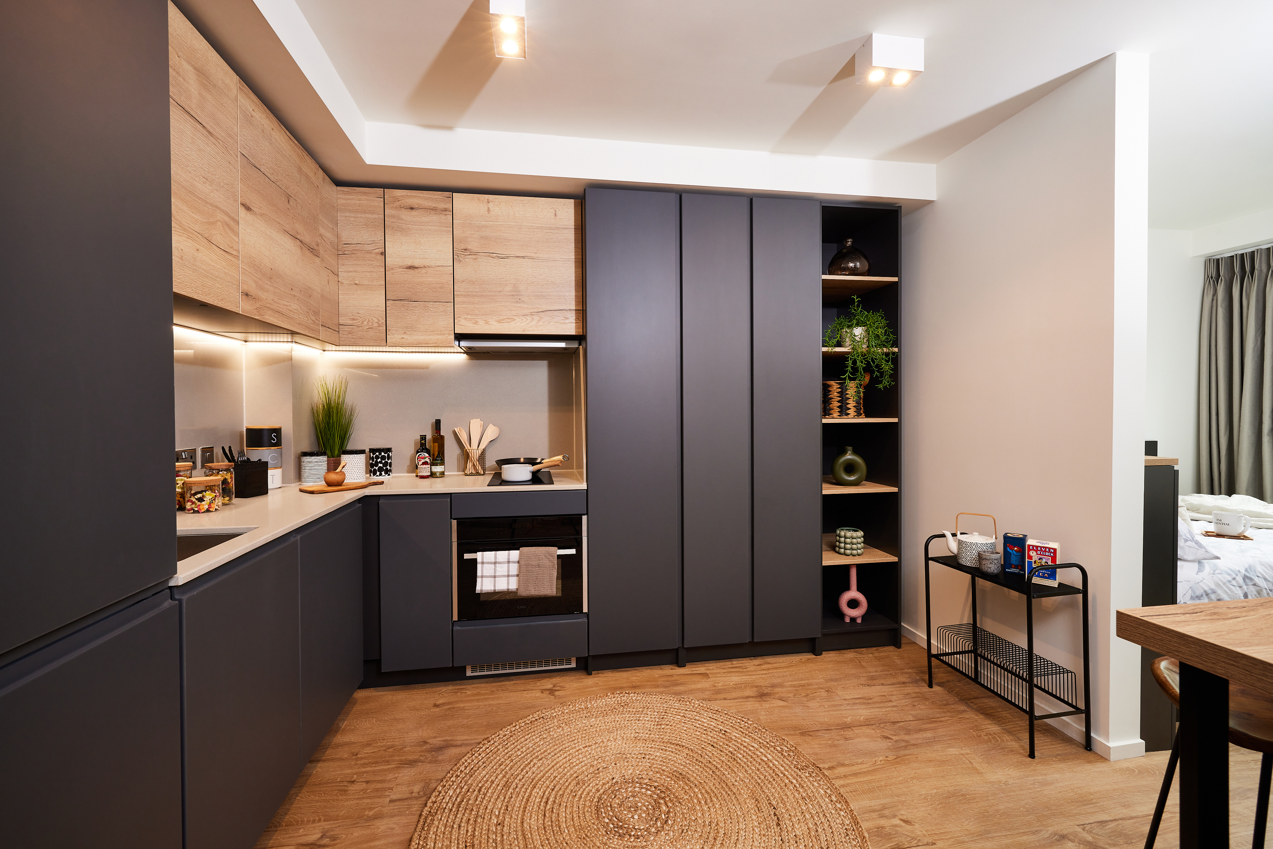 Modern kitchen with matte black cabinets and wood accents, inspired by Cannon Park's sleek design. Features an oven, sink, open shelving with decor, and a rug on wood flooring. A small table with snacks and a cozy lit area add Warwick charm in the background.