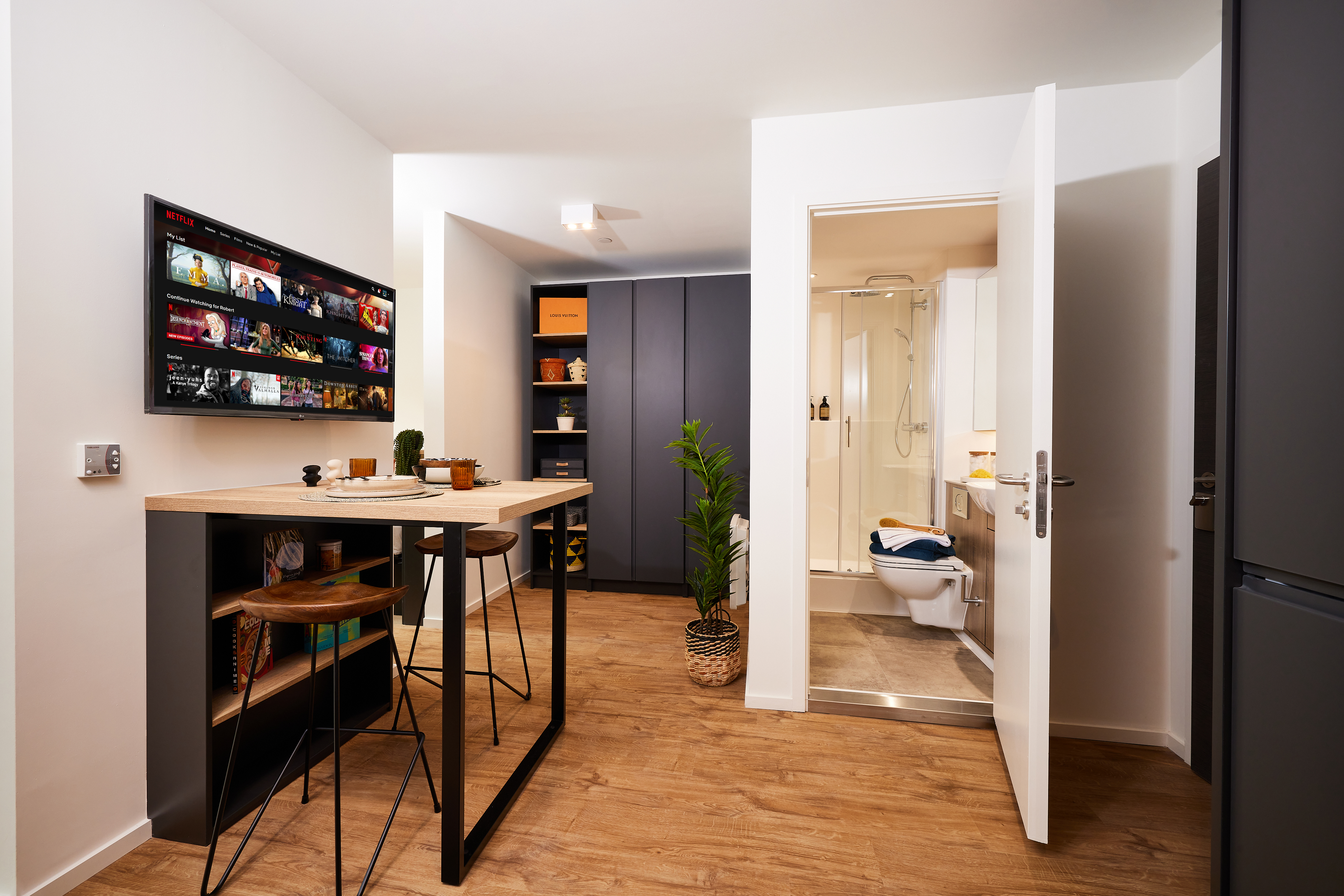 Nestled in the heart of Warwick, this modern studio apartment features wooden flooring, a small kitchen island with two stools, and a wall-mounted TV displaying colorful images. The room boasts black cabinets, a plant, and a doorway leading to a bathroom with a sleek glass shower.