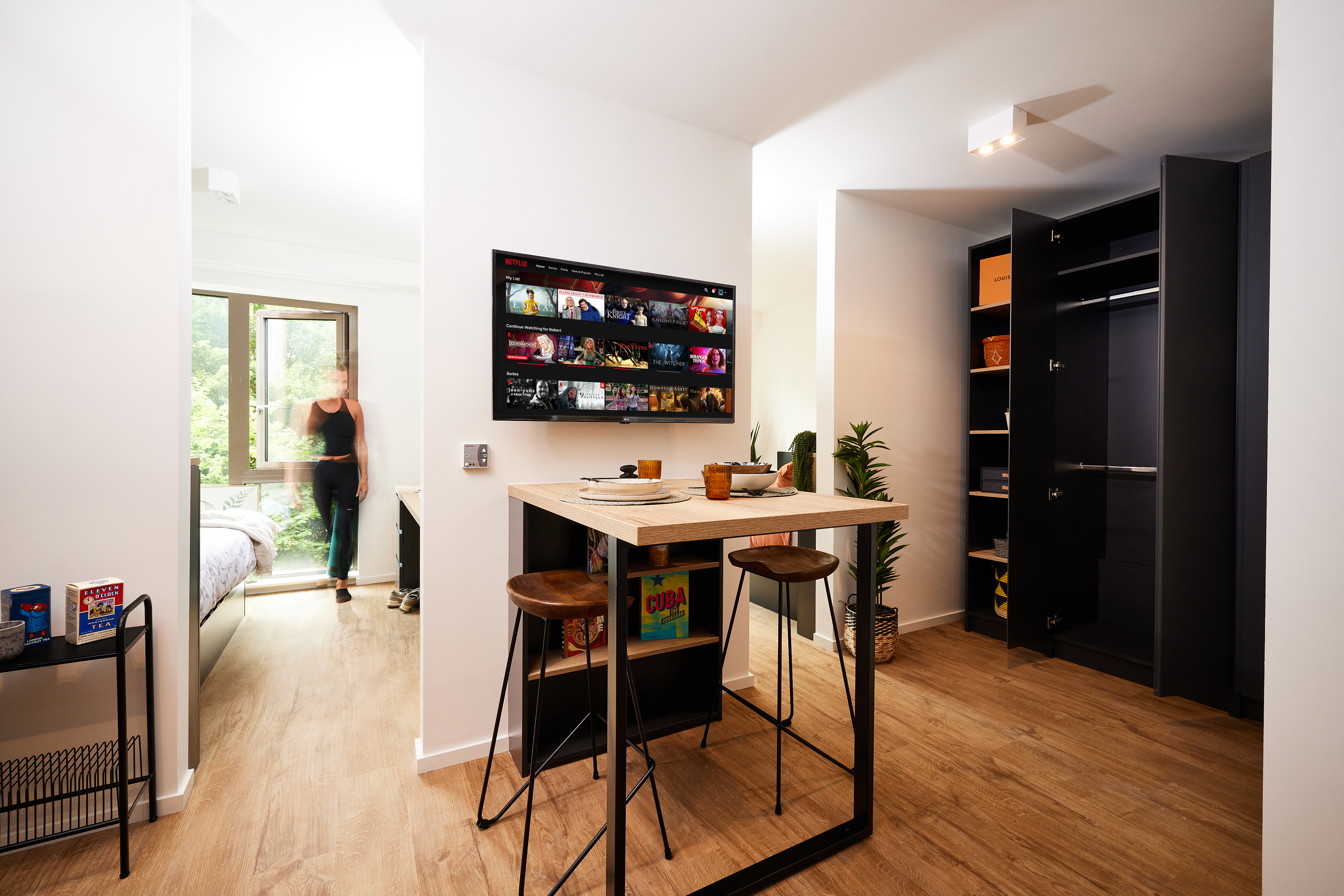 Modern apartment interior in Cannon Park with a small kitchen and dining area featuring a high table and two stools. A person is seen near a bed in an adjacent room. A wall-mounted TV displays streaming options. Wooden flooring and a partially open closet are visible, embodying Warwick's charm.