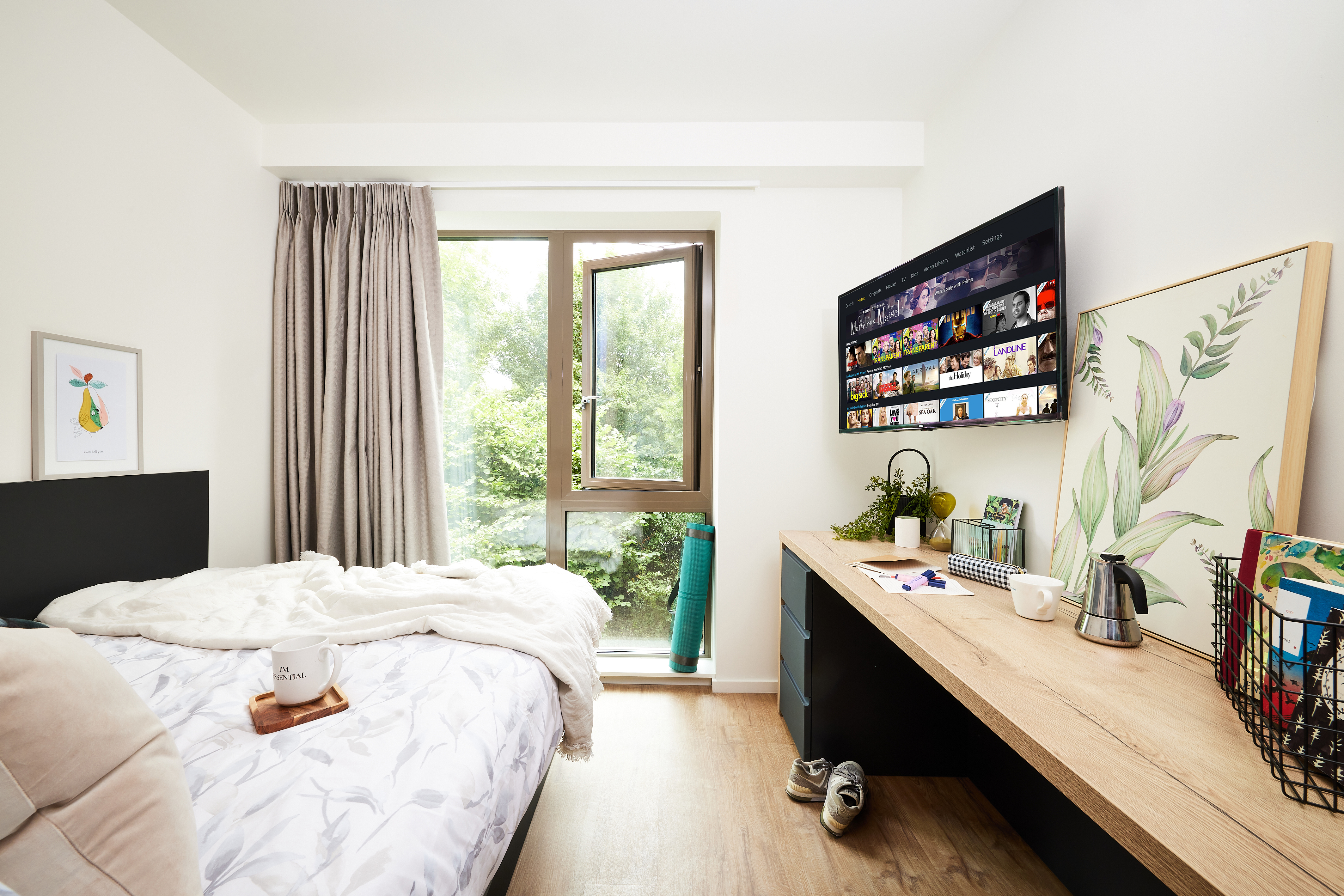 A tidy bedroom with a bed, gray curtains, and an open window overlooking the greenery of Cannon Park. A desk holds a kettle, plants, and a wire basket. A wall-mounted TV displays colorful images of Warwick, while a yoga mat rests by the window. Artwork decorates the walls.