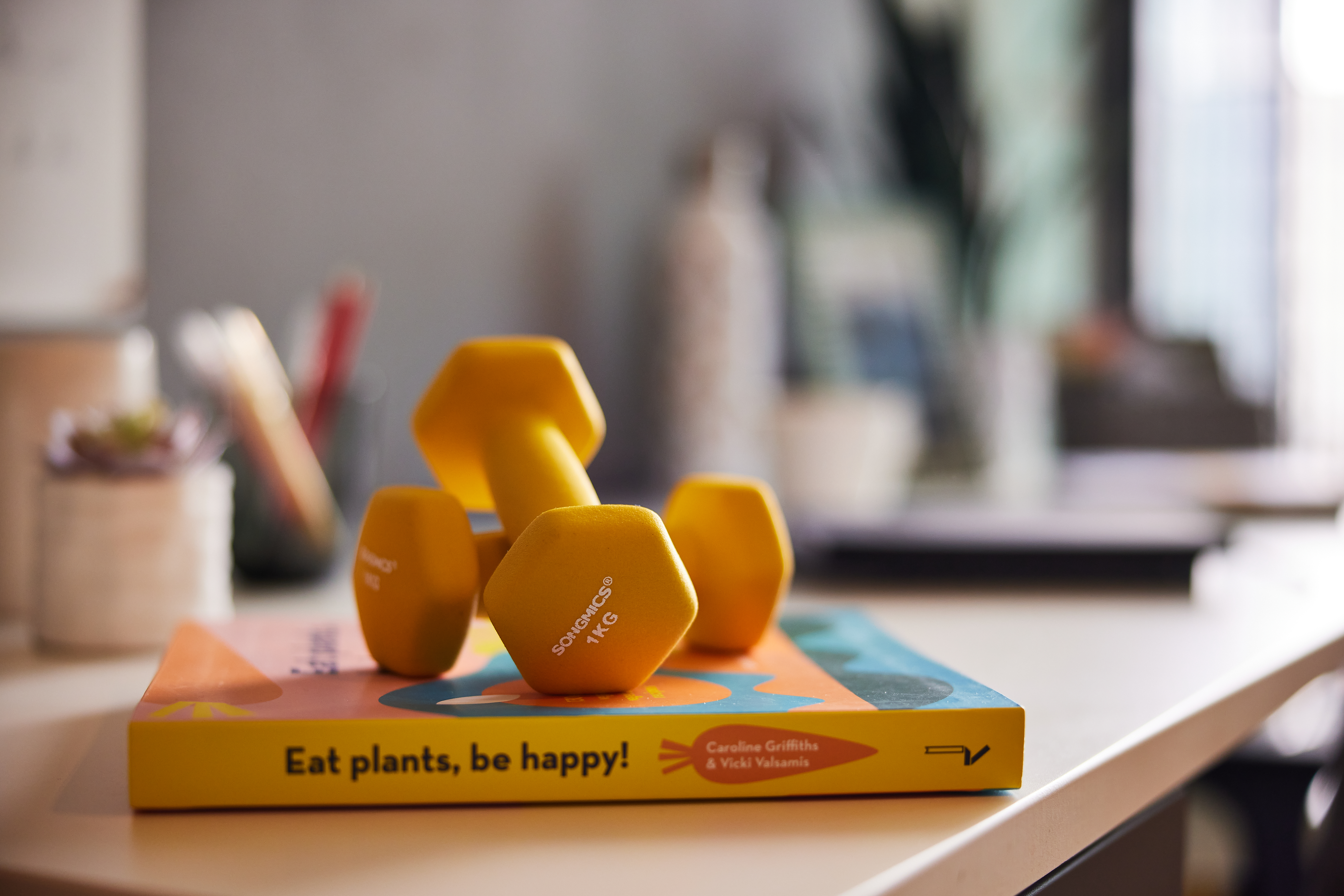 A pair of yellow 1 kg dumbbells rests on a colorful book titled "Eat plants, be happy!" The background evokes the vibe of student accommodation, with its blurred interior filled with plants and various desk items, suggesting a cozy, bright atmosphere.