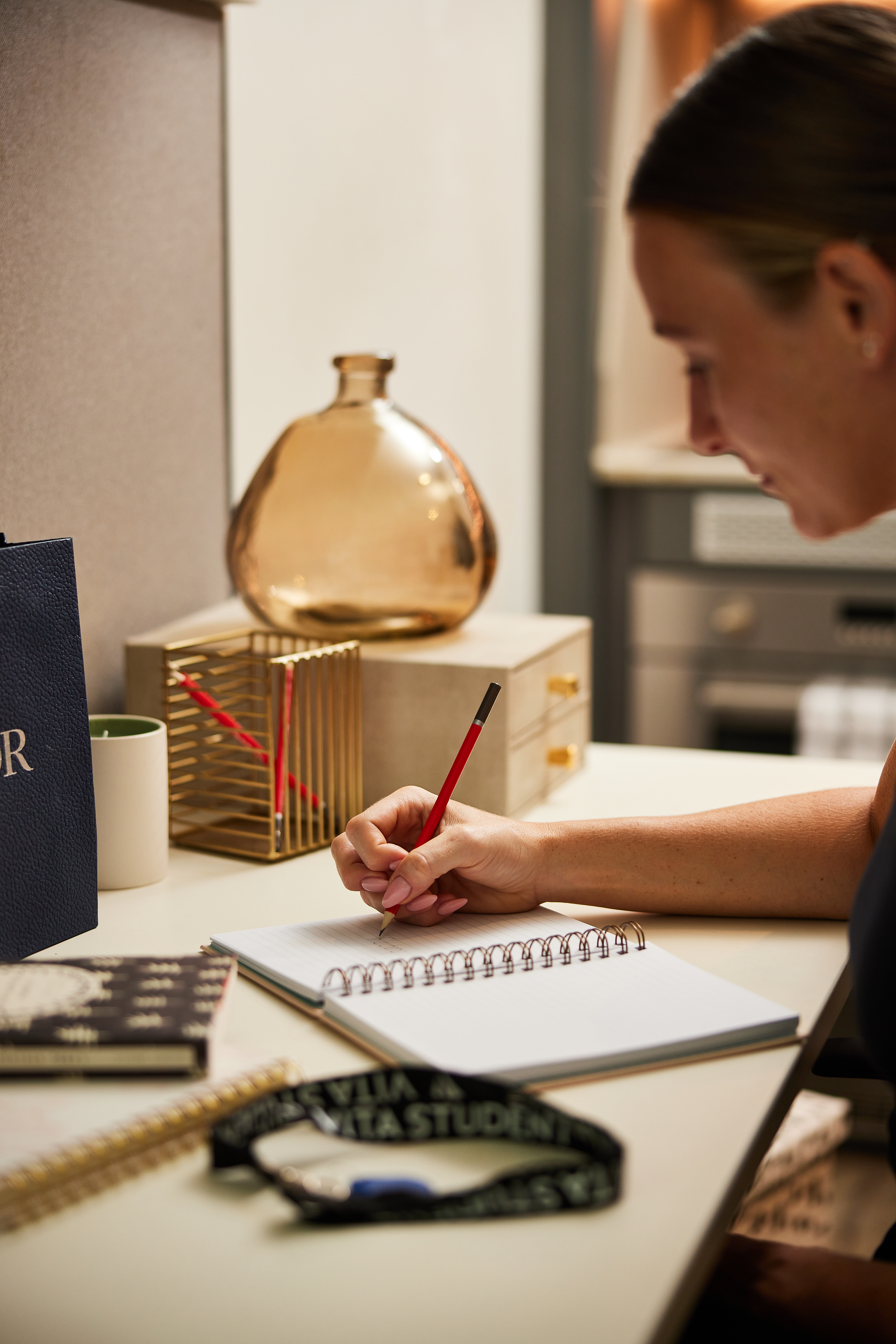 A person writes in a notebook at a desk with a red pencil. The cozy Portland House setting includes a gold wire organizer, a glass vase, notebooks, and a lanyard on the desk. In the background, there's a cabinet and cup adding to the ambiance.