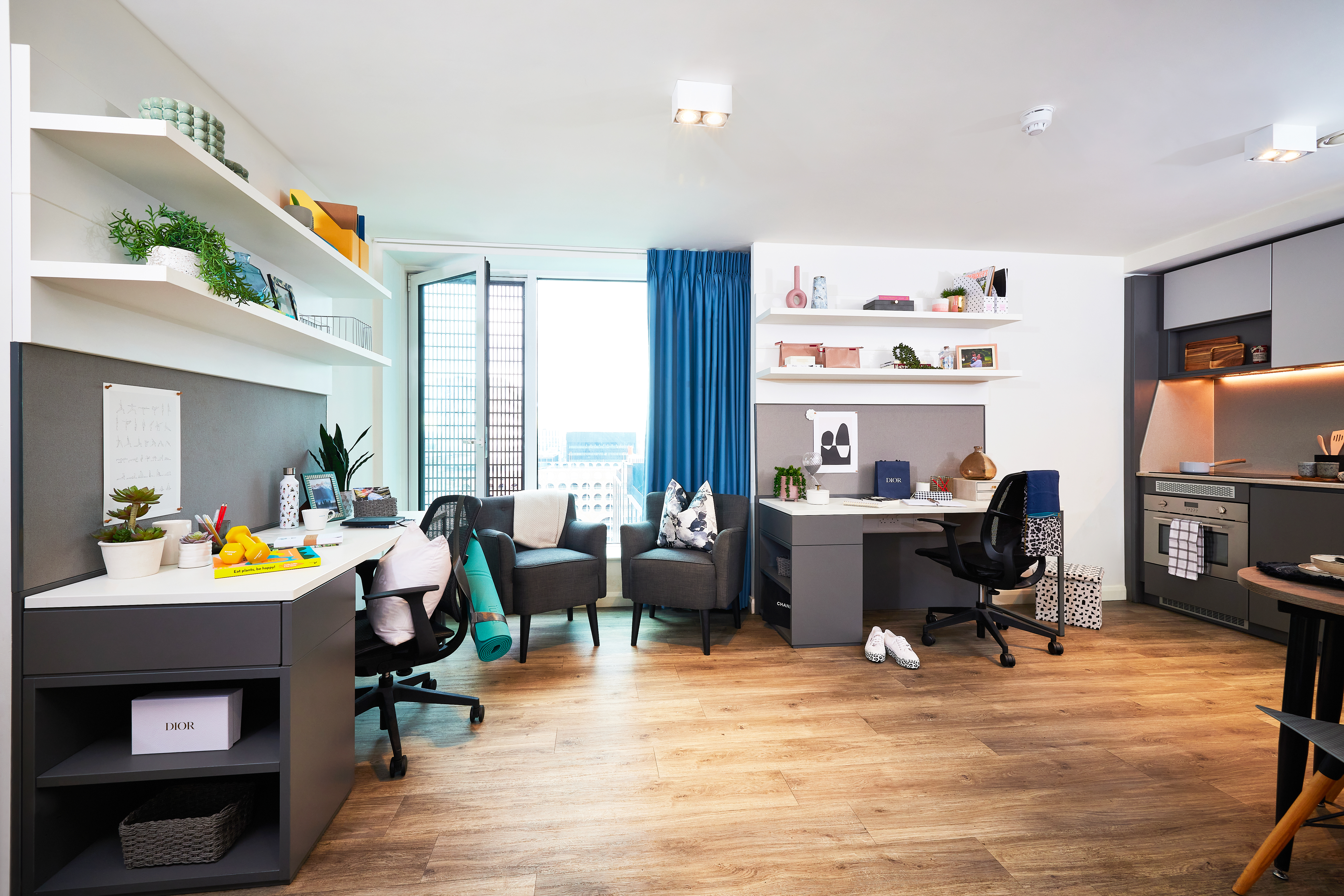 Bright, modern studio apartment at Circle Square with wood flooring. Features two desks with chairs, bookshelves, plants, and personal items. The space includes a cozy sitting area, small kitchen, and an open window with blue curtains offering a city view.