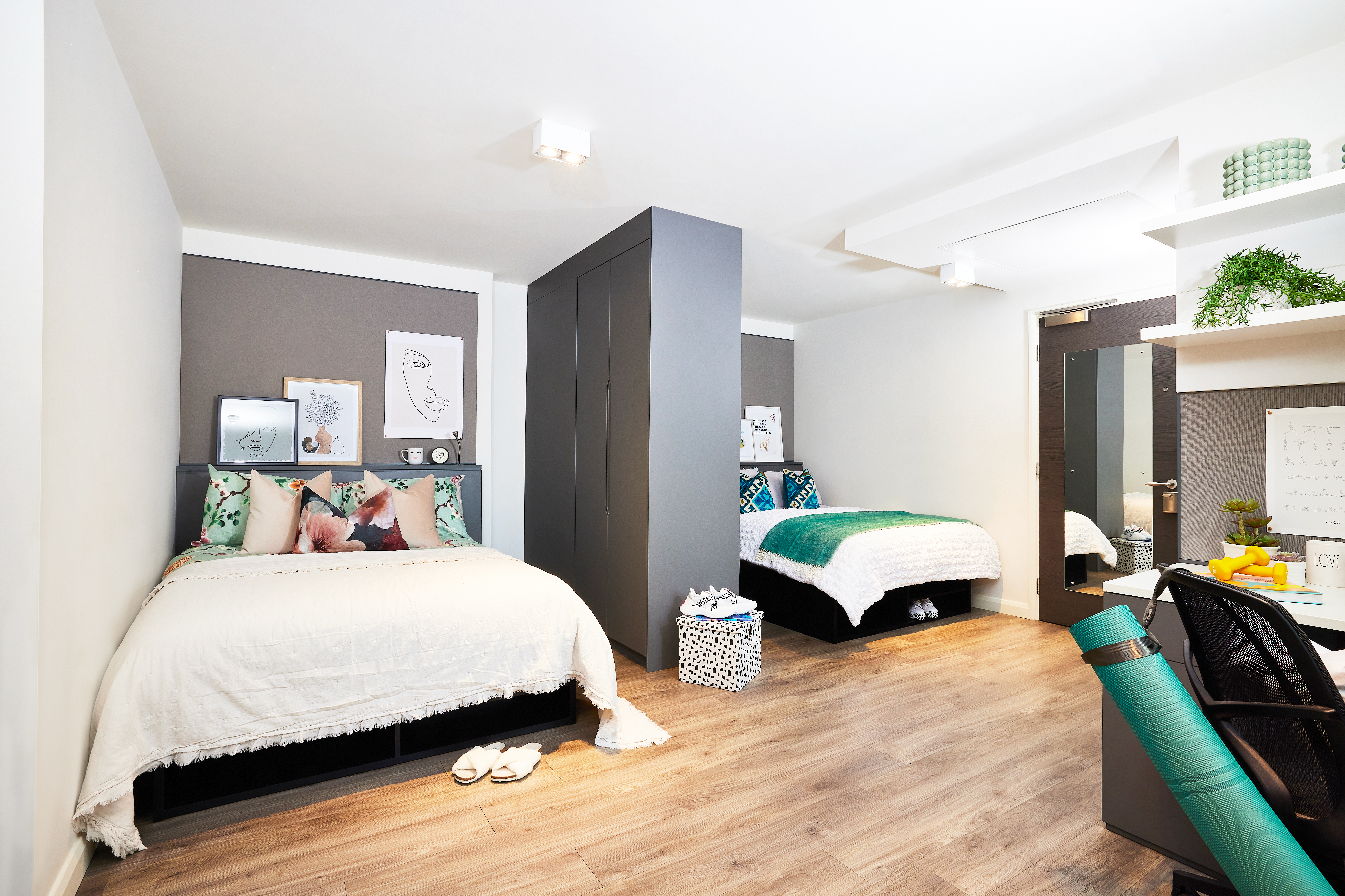 A modern, spacious bedroom features two neatly made beds with decorative pillows. A central wardrobe separates the beds, alongside square-patterned wooden flooring. A small desk, chair, and plants enrich the cozy and organized ambiance.