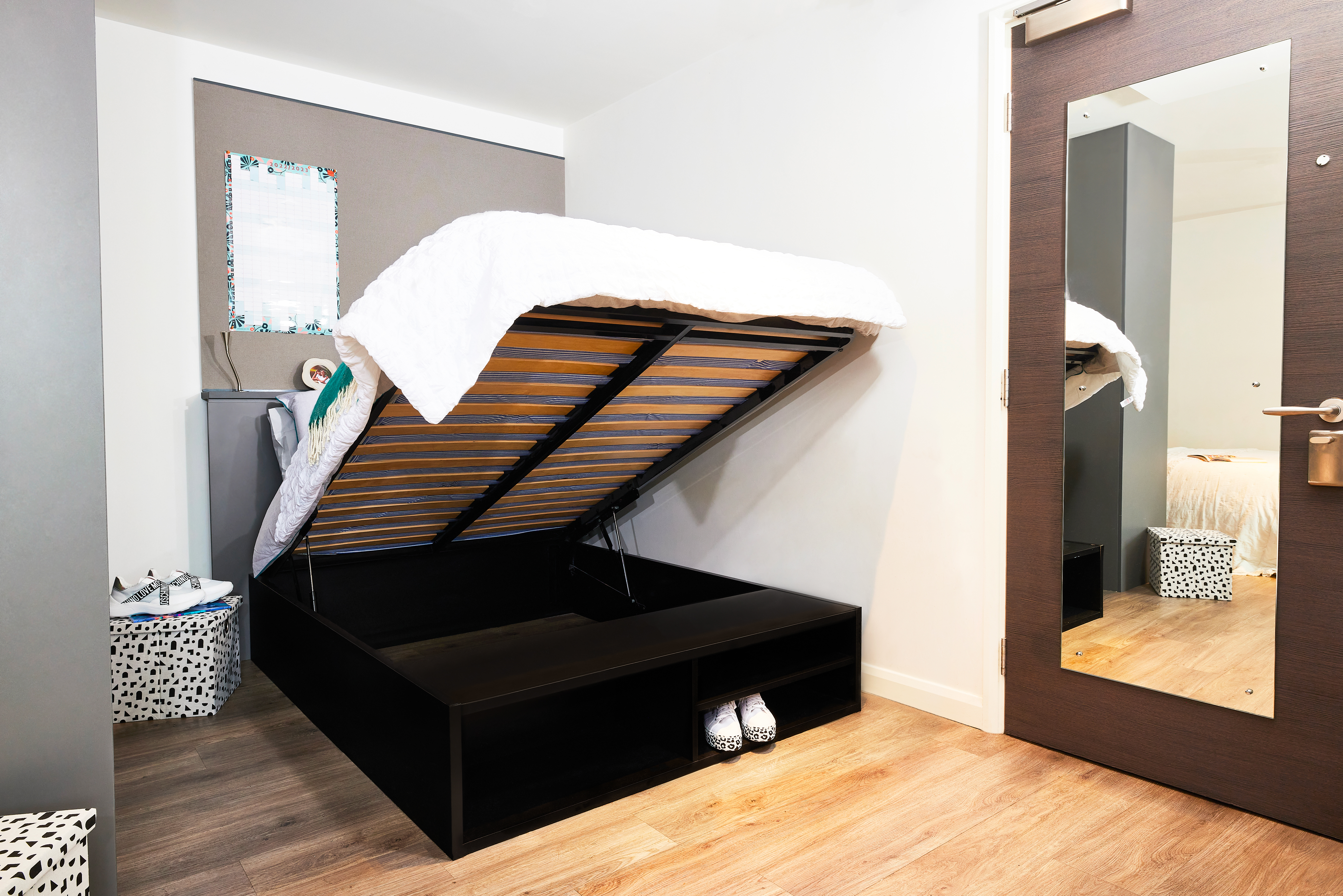 A modern bedroom features a hydraulic bed with its mattress lifted, revealing storage space below. The room includes a full-length mirror on the door, a laundry basket, and a minimalist decor style with square wooden flooring.