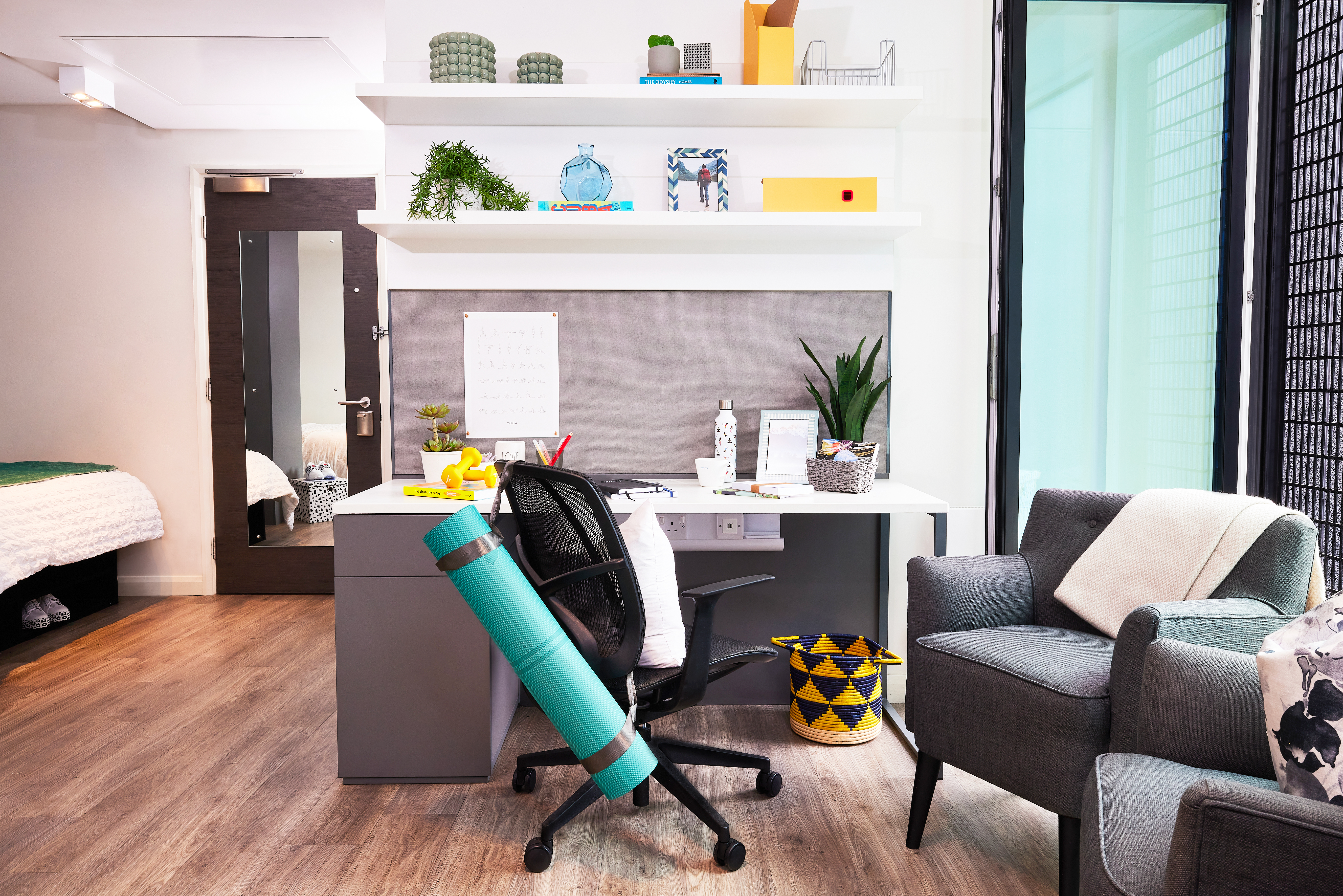 A modern home office features a square desk and ergonomic chair, with a yoga mat leaning against it. Shelves above hold plants, books, and decorative items. Two circular gray armchairs sit in the corner, while a bed is partially visible in the adjoining room.