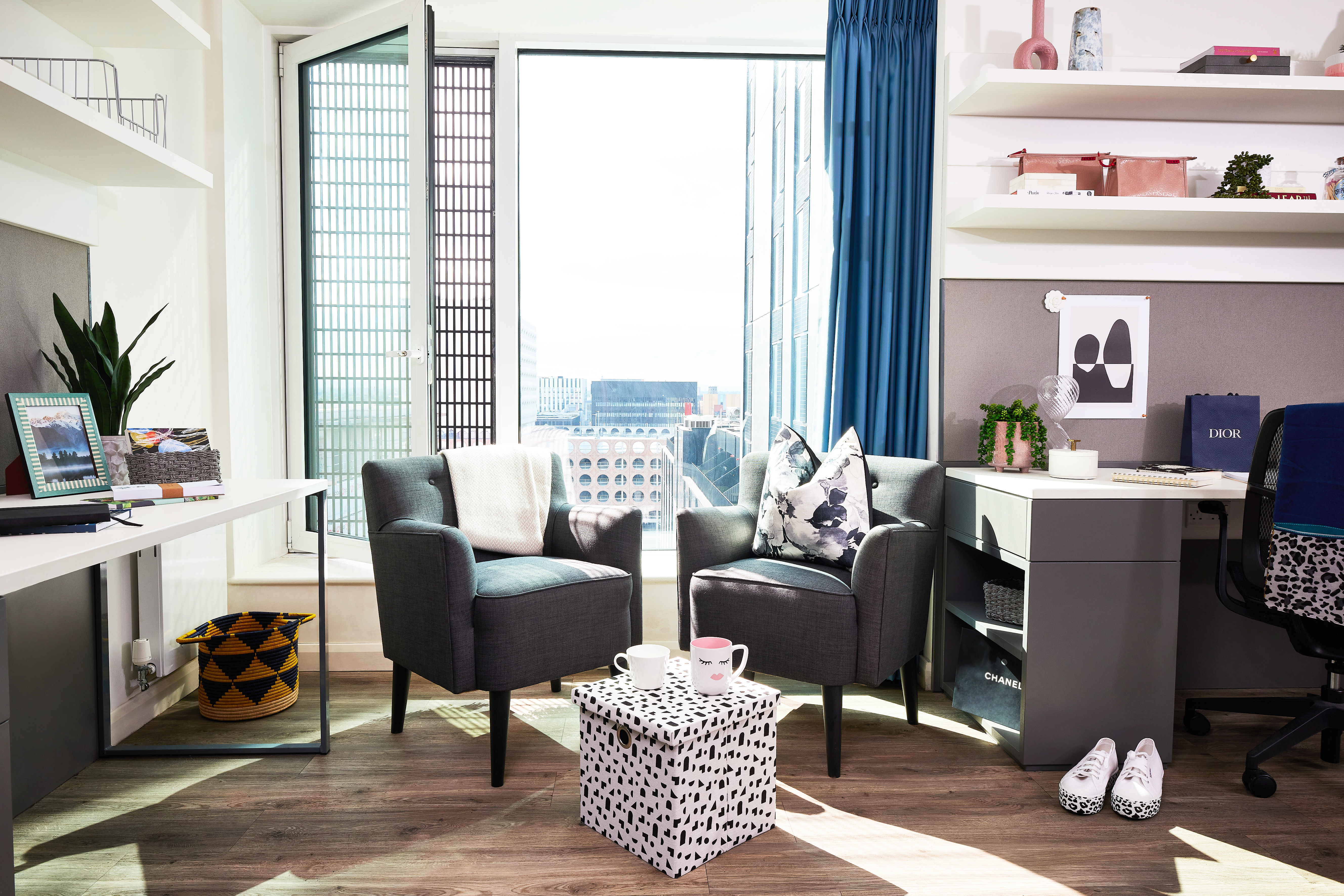A modern office space features two gray armchairs with patterned pillows, a small round table with mugs, and a floor-to-ceiling window revealing a city view. A desk holds a computer, plant, and storage squarely organized, while shelves display decor items.