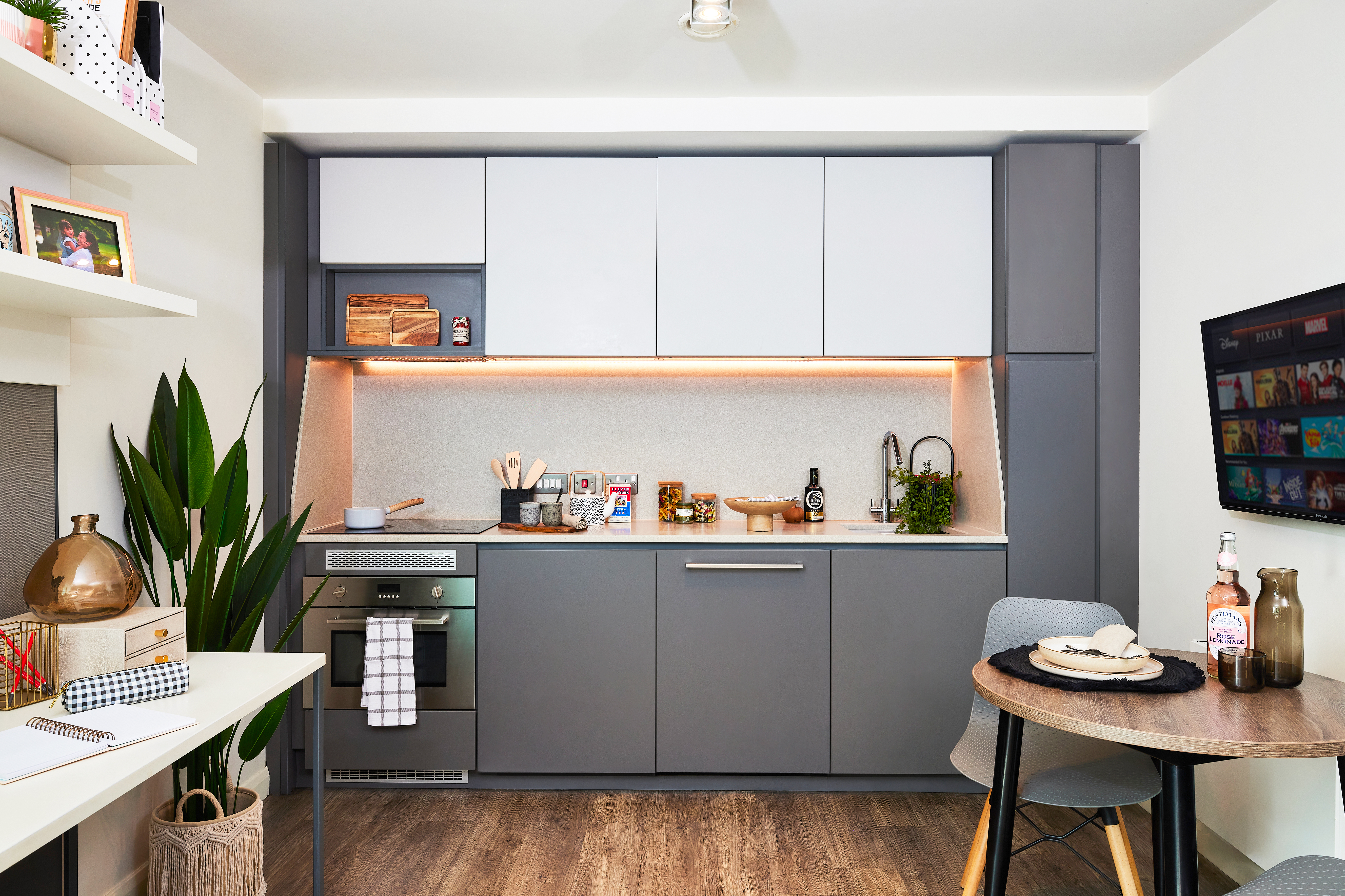 A modern kitchen with gray cabinets, a built-in oven, and a small wooden dining table sits elegantly under a square window. Plants and kitchen items decorate the space. A TV is mounted on the wall, and light streams in, illuminating the wooden floor.