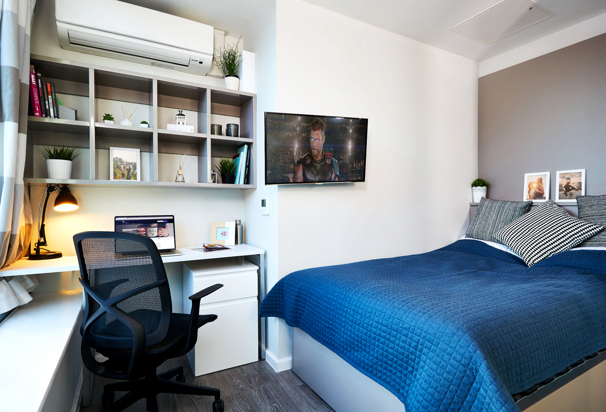 The modern bedroom in this luxury student accommodation features a neatly made bed with a blue quilt, a wall-mounted TV, and a desk with a laptop and lamp. Shelves hold books and plants beneath efficient air conditioning, keeping the bright room tidy and comfortable.