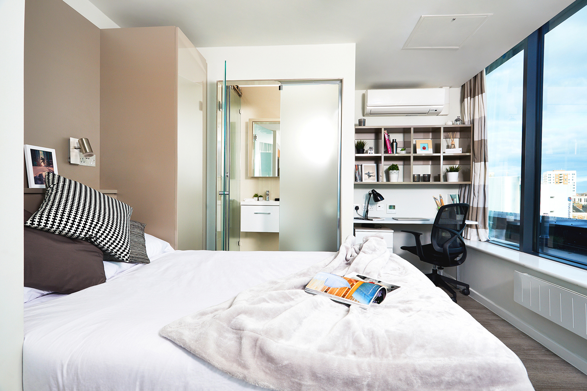 A luxury student accommodation offers a modern bedroom with a neatly made bed, cushions, and a gray throw blanket. It features a glass-walled bathroom, a desk with lamp and books, and large windows with city views. The room boasts minimalist decor and an air conditioner for ultimate comfort.