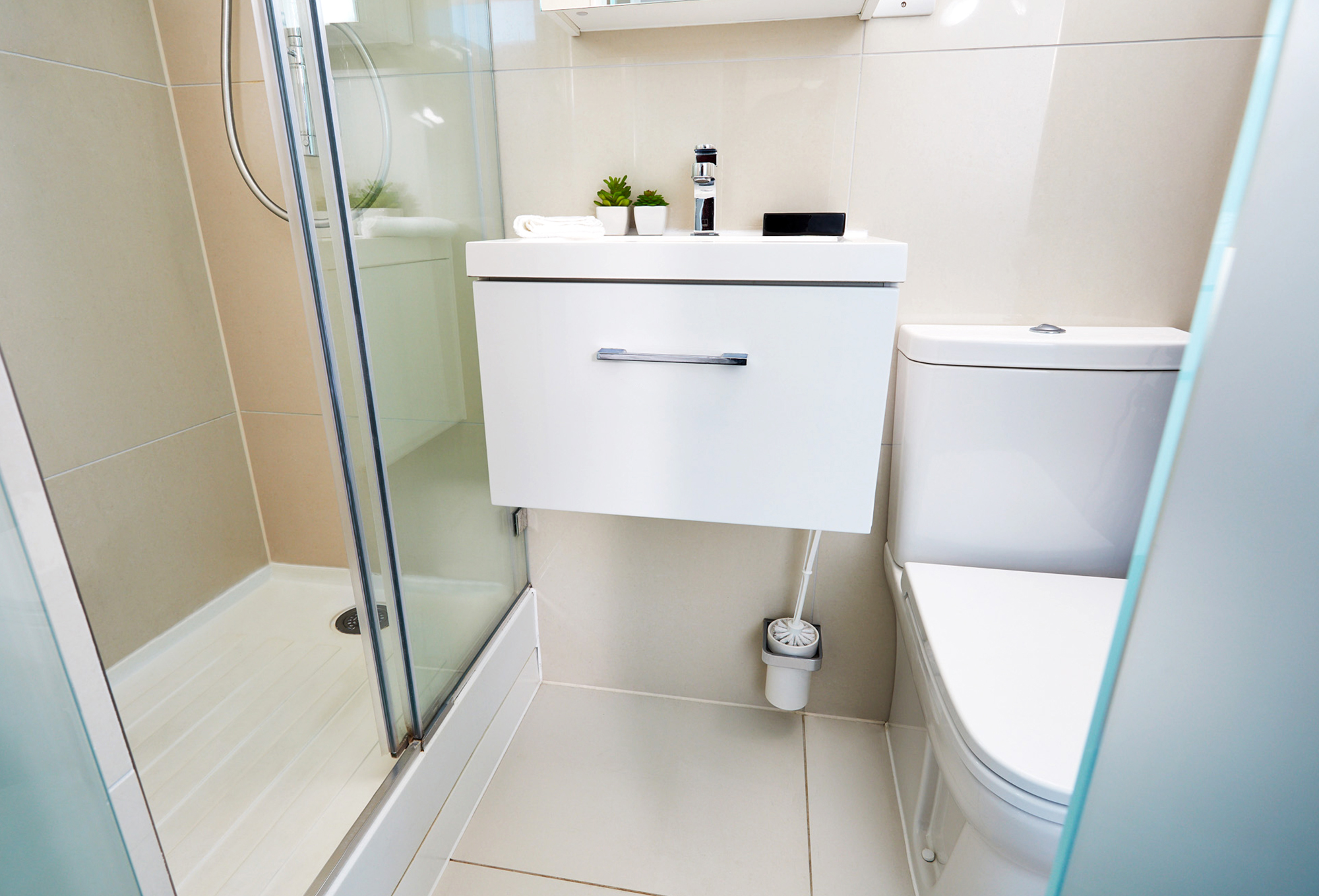 This modern bathroom in the luxury student accommodation features a glass shower enclosure on the left, a wall-mounted white sink with a sleek metal faucet and two small potted plants, and a white toilet on the right. Light-colored tiles and minimal decor complete this elegant space.