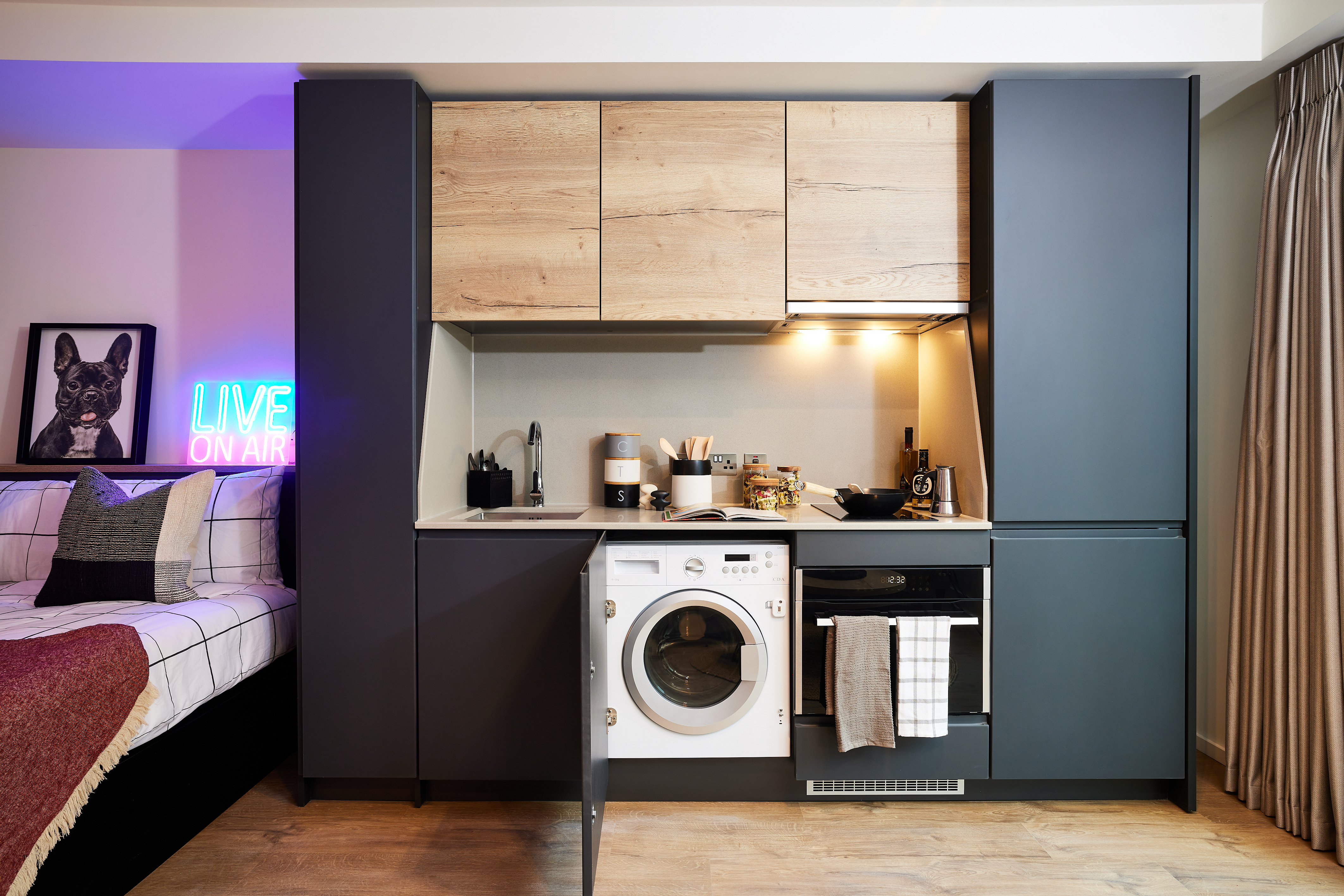 Compact kitchen with dark cabinets, featuring a washer, oven, and open cabinet doors revealing appliances. Above, wooden wall units. Adjacent bed with plaid bedding. Neon "LIVE ON AIR" sign and bulldog artwork add flair. Curtains to the right complete this Warwick-inspired space near Cannon Park.