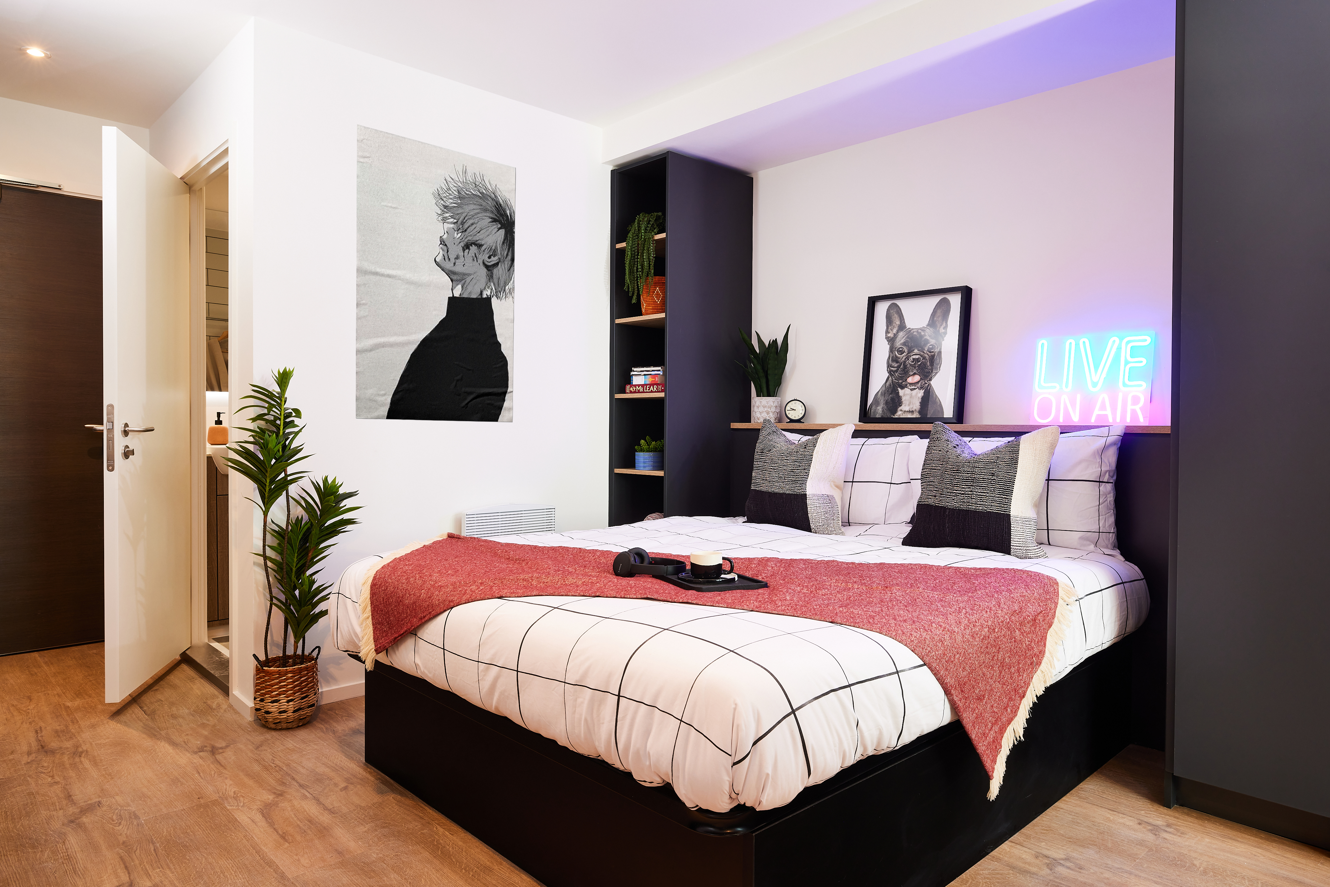 Modern bedroom in Cannon Park with a neatly made bed featuring a red blanket and grid-patterned bedding. A neon sign reads "LIVE ON AIR" above the bed. A black and white dog portrait is displayed. The room includes plants and an open door leading to a Warwick-style bathroom.