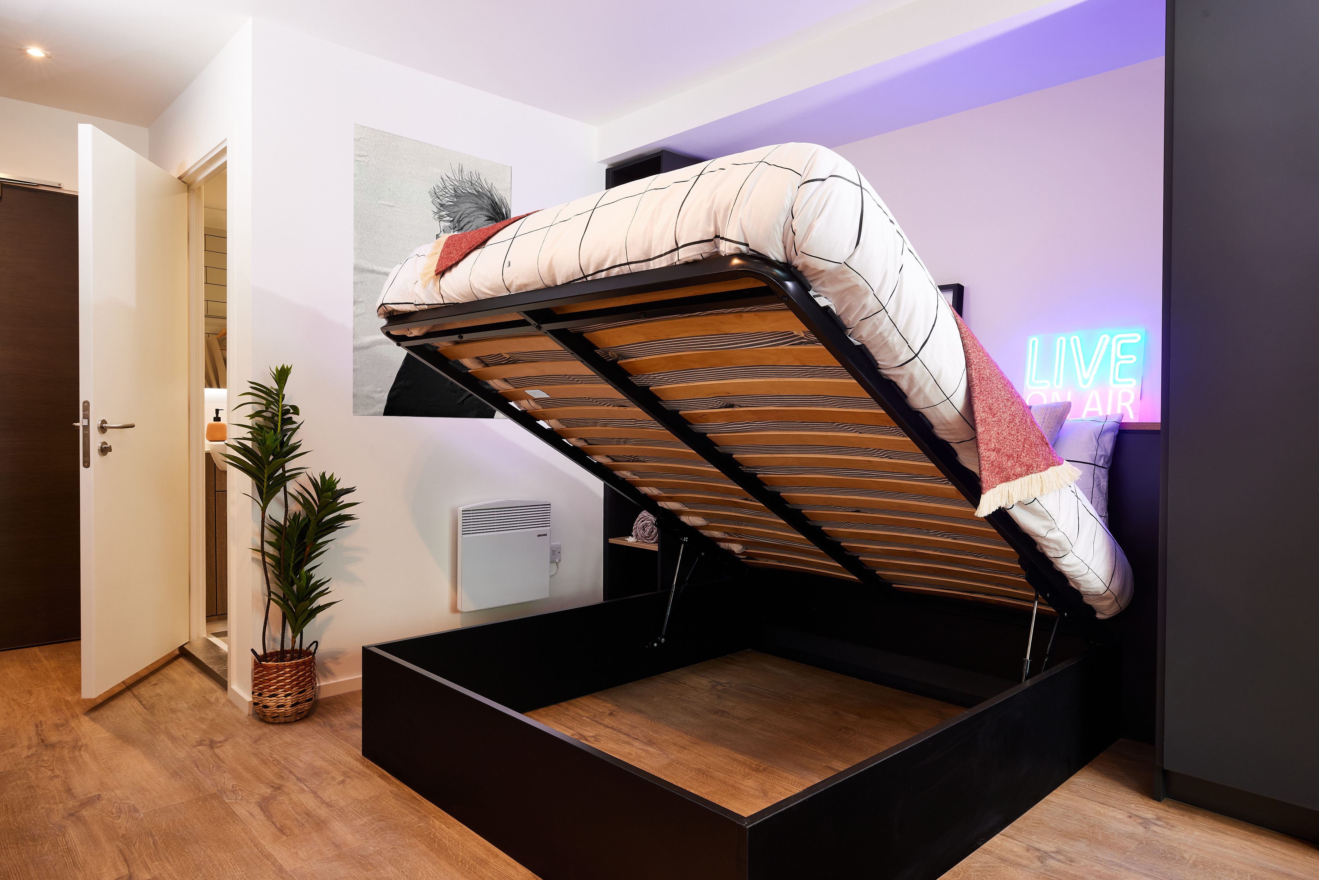 A modern room in the Warwick style features a bed with a lifted mattress revealing storage underneath. A neon sign on the wall reads "Live on Air." A plant sits in the corner, and there's an open door leading to another room, reminiscent of sleek spaces in Cannon Park.