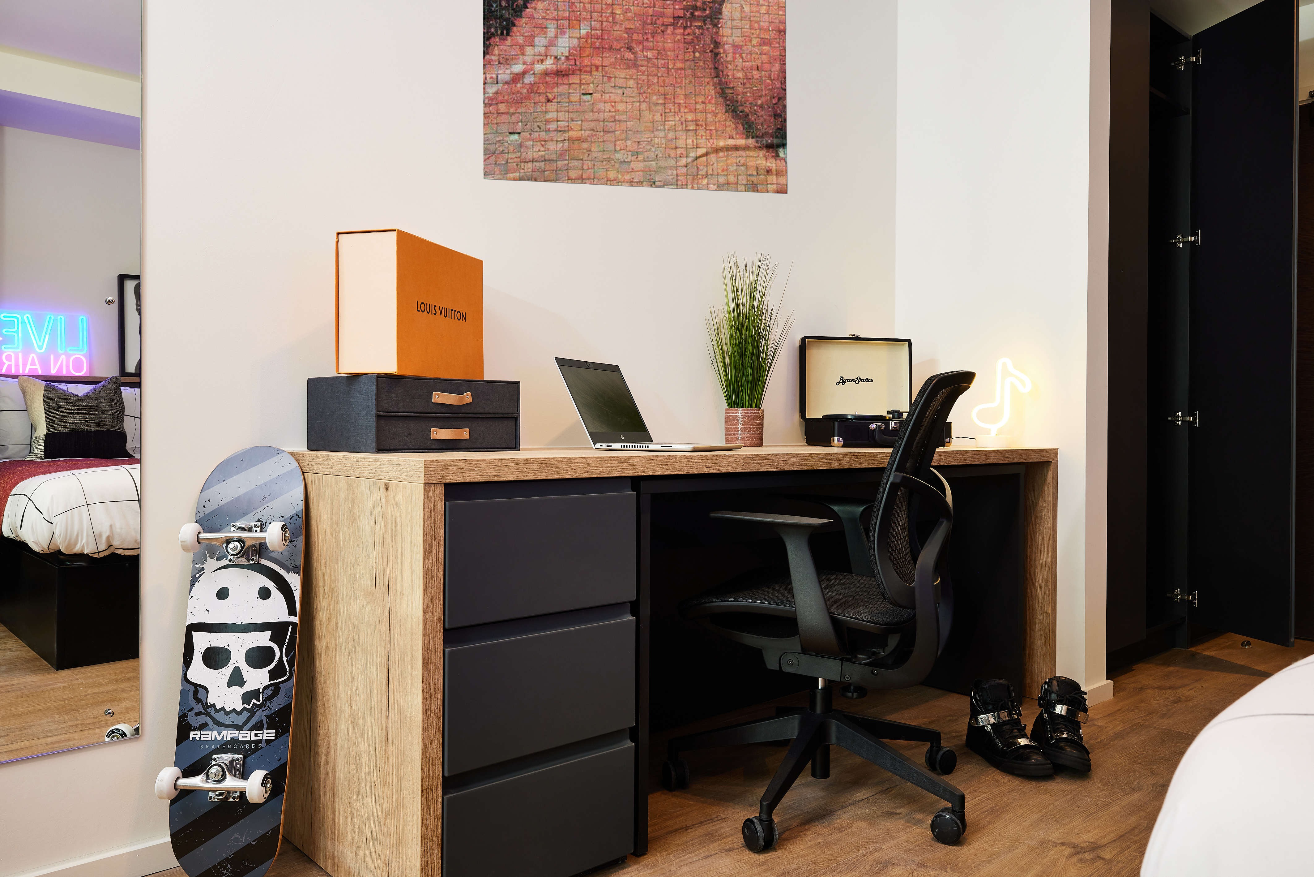 A modern bedroom in Warwick features a wooden desk and black chair. Items on the desk include a laptop, books, a potted plant, and a neon flamingo light. A skateboard leans against the desk. The bed has a colorful pillow, and artwork hangs above it, reminiscent of Cannon Park's vibrant scene.