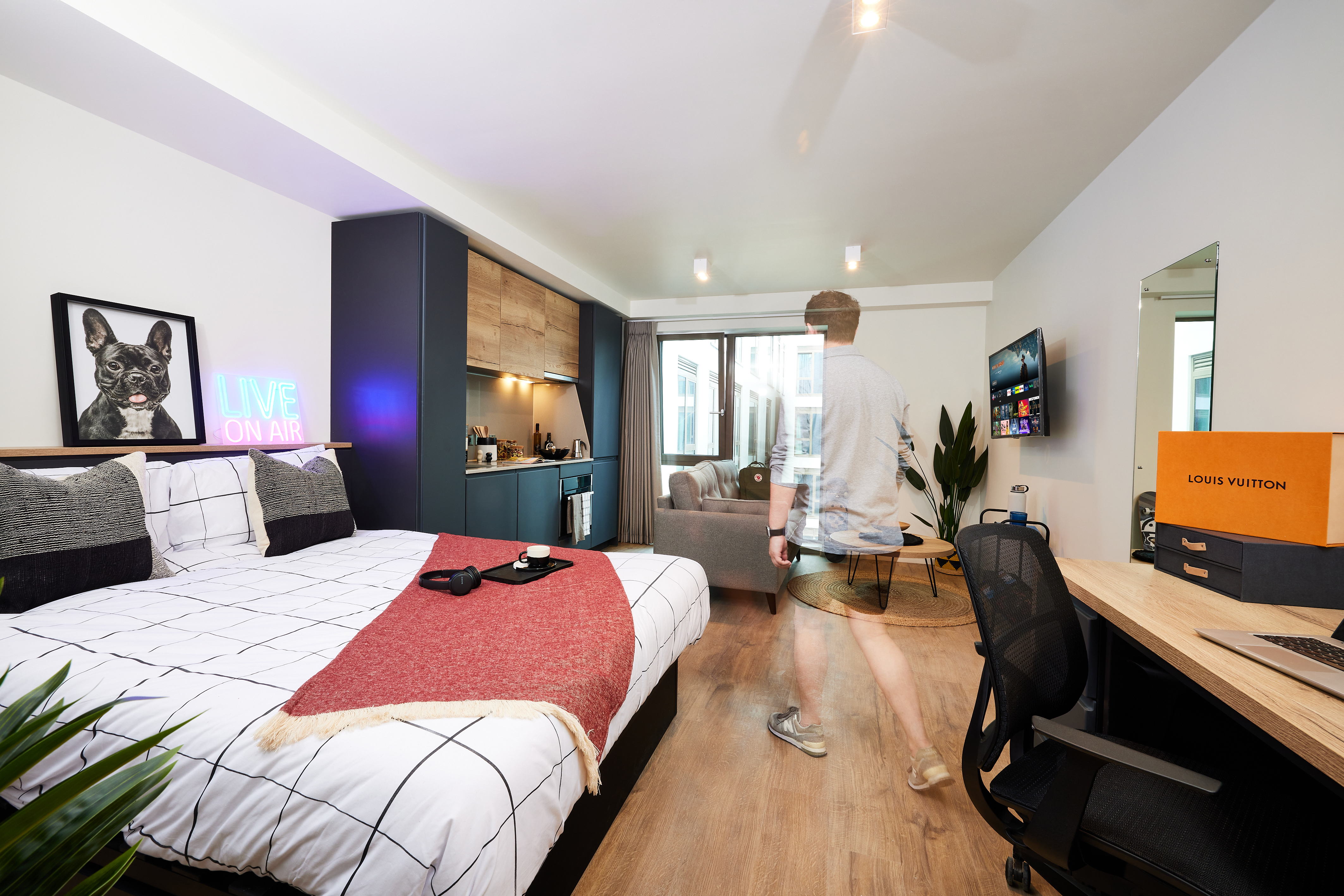 A modern studio apartment with a bed, desk, and kitchen area. A blurred person walks by, showcasing decor including a "LIVE ON AIR" sign and luxury brand boxes. Neutral tones dominate, with greenery and natural light from large windows completing the space.