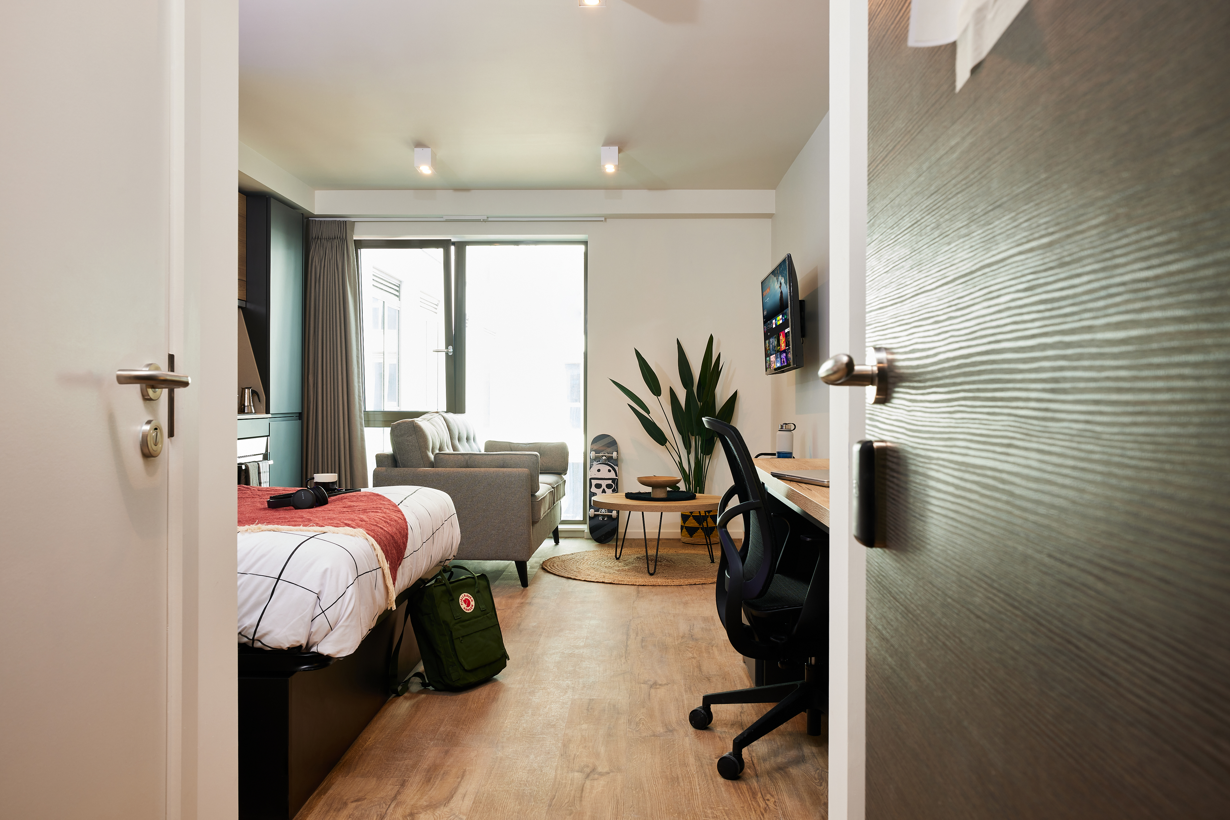 Peering through the open door, this modern dorm room at Warwick sports a bed with striped bedding and a red blanket. On the right, a desk with a computer and an office chair awaits, while sunlight filters through a large plant by the window. Nearby, a skateboard rests against the wall, reminiscent of Cannon Park's vibe.