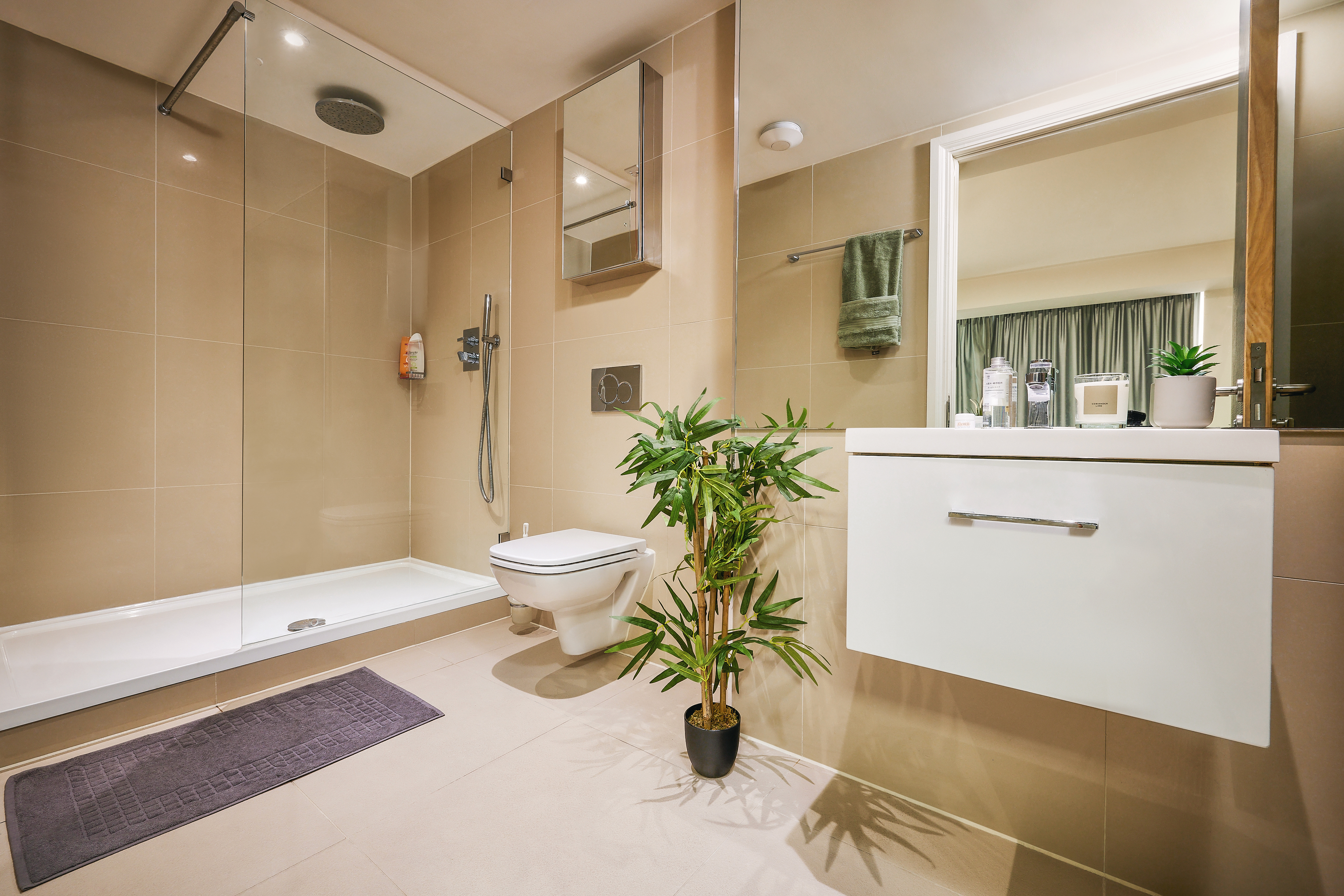 Modern bathroom with a glass-enclosed shower, wall-mounted toilet, and a white vanity with a mirror above. A green plant rests by the vanity, adding a touch of nature to the house. Soft lighting brightens the beige-tiled walls and floor.