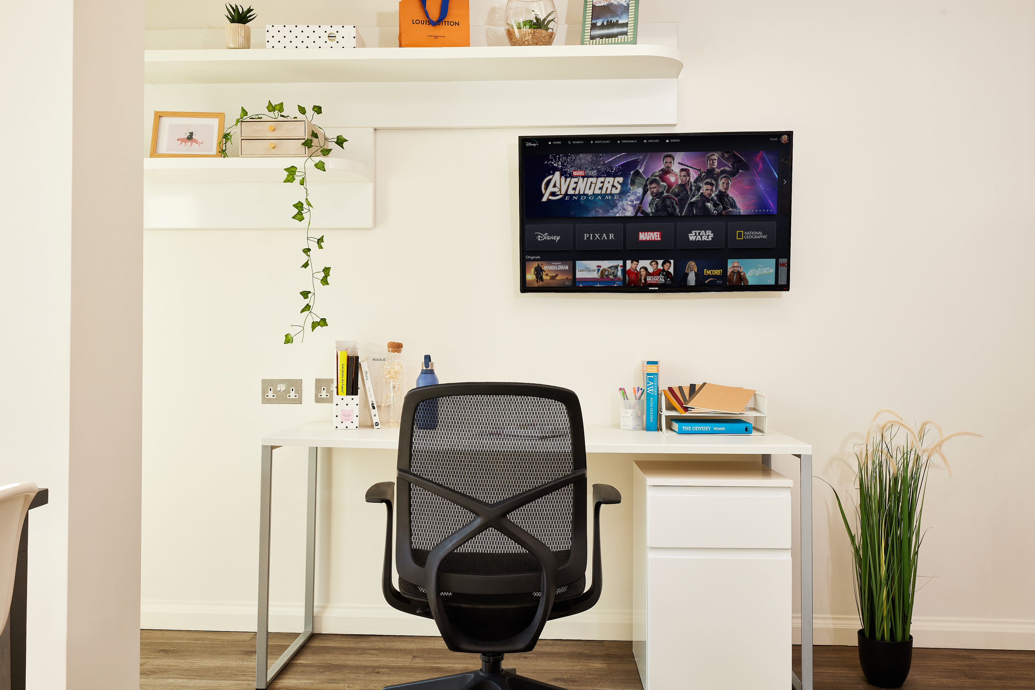 Modern home office with a sleek desk and mesh chair facing a wall-mounted TV displaying a streaming service. Shelves above boast plants and decor. A vintage telephone on the cabinet adds charm, while light-colored walls and flooring enhance the room's airy ambiance, complemented by a potted plant to the right.