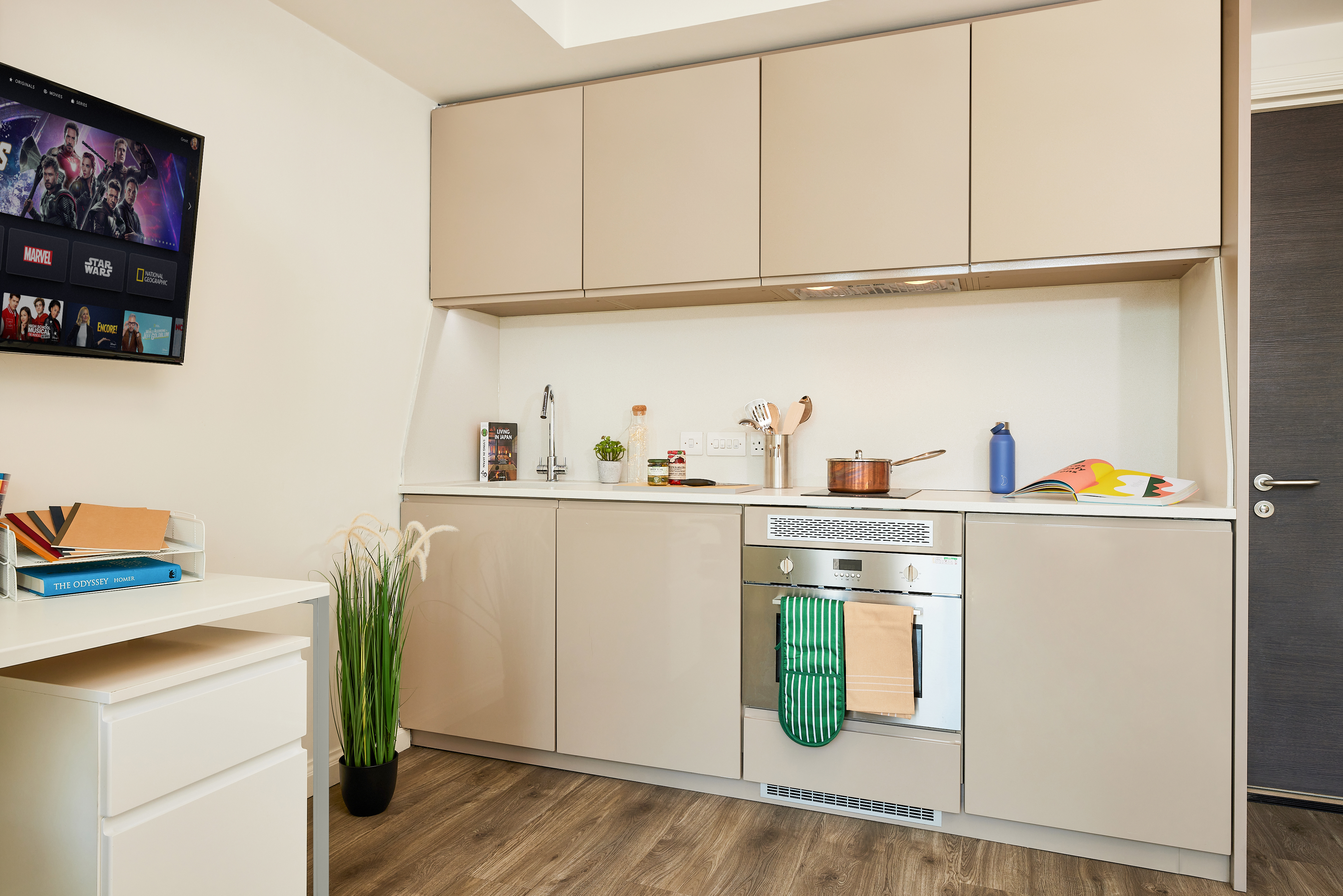 A modern kitchen showcases beige cabinets and an oven with towels hanging, while a countertop holds a kettle, jar, and open cookbook. A TV, streaming content, is mounted on the wall. Nearby furniture features a plant, books, and a vintage telephone for a touch of timeless charm.